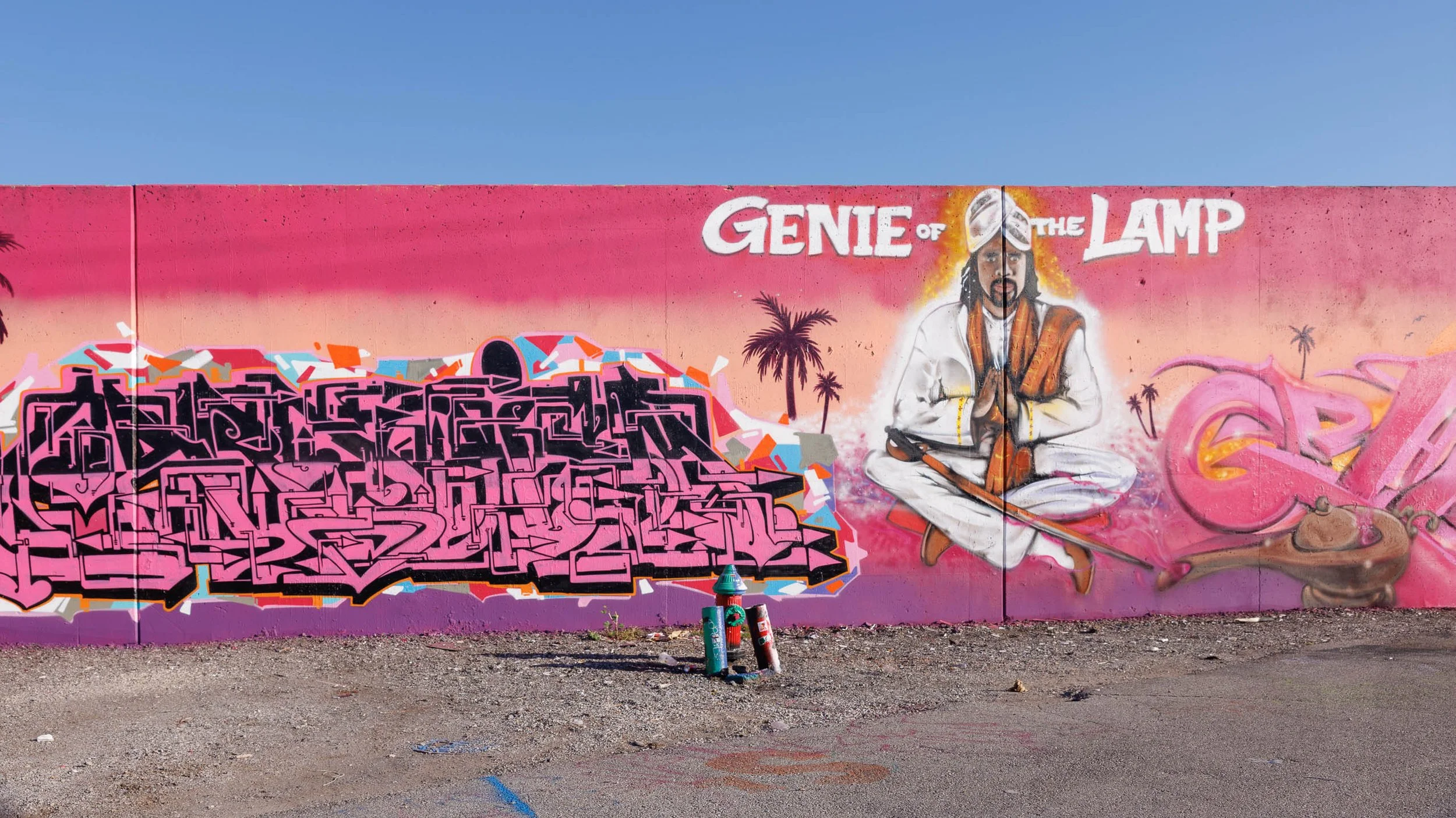 Long magenta-to-peach mural wall in St. Louis with pink graffiti lettering at left, silhouetted palm trees, a seated white-robed figure under the words Genie of the Lamp and a brown lamp at right