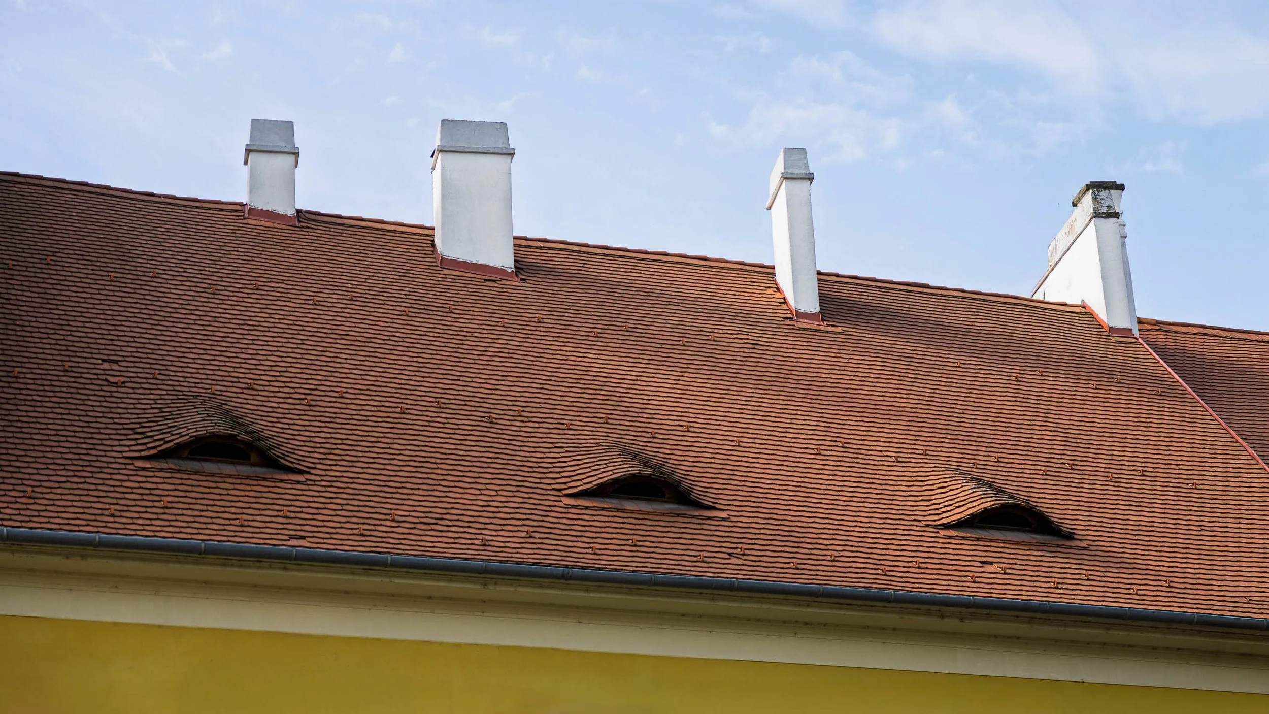 Three eye-shaped dormer windows and four white chimneys punctuate a terracotta roof beneath a pale blue sky in Prague, Czech Republic.