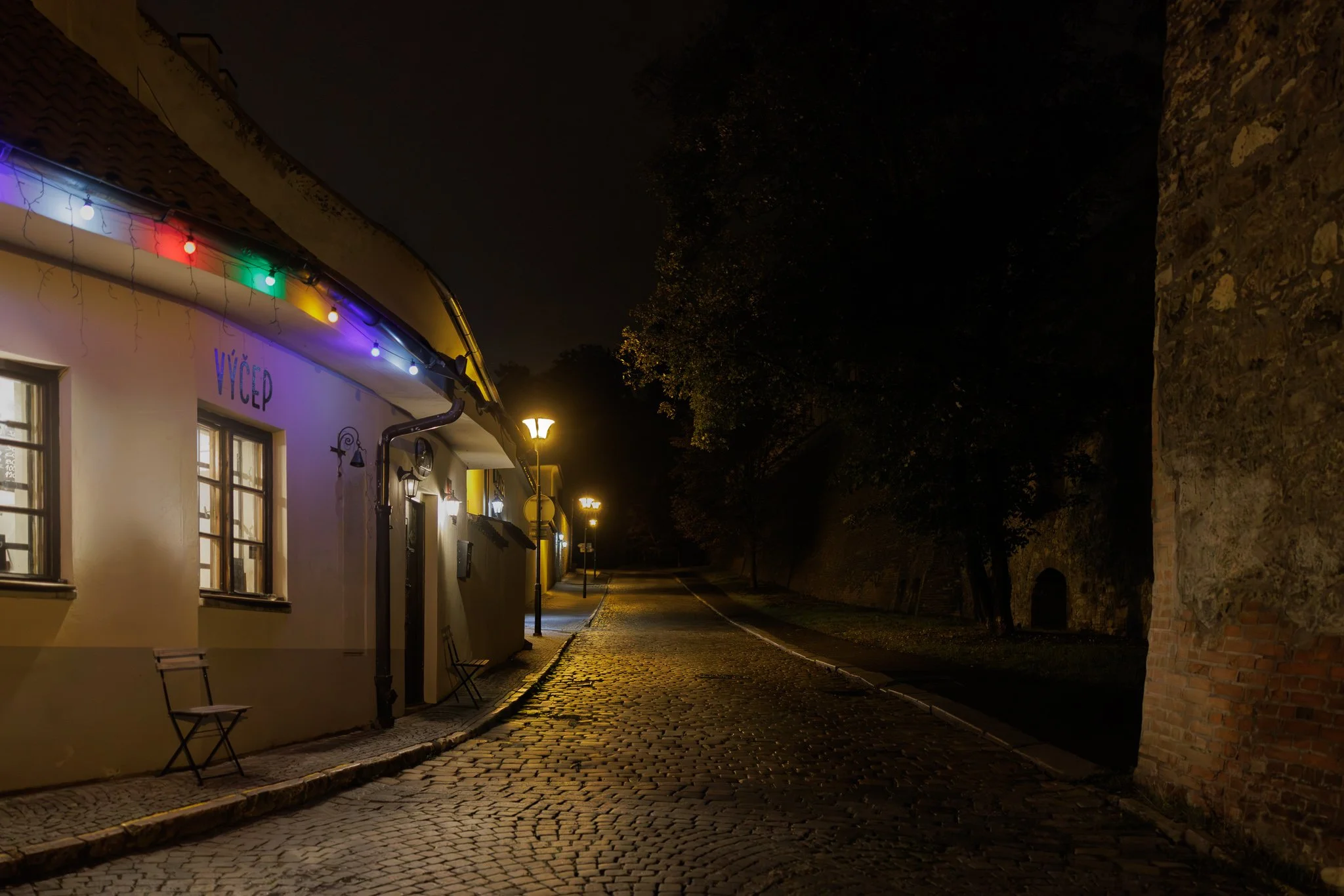 Quiet Prague street at night with colored café lights, empty chairs, warm streetlamps, cobblestone pavement, and historic stone walls fading into darkness.