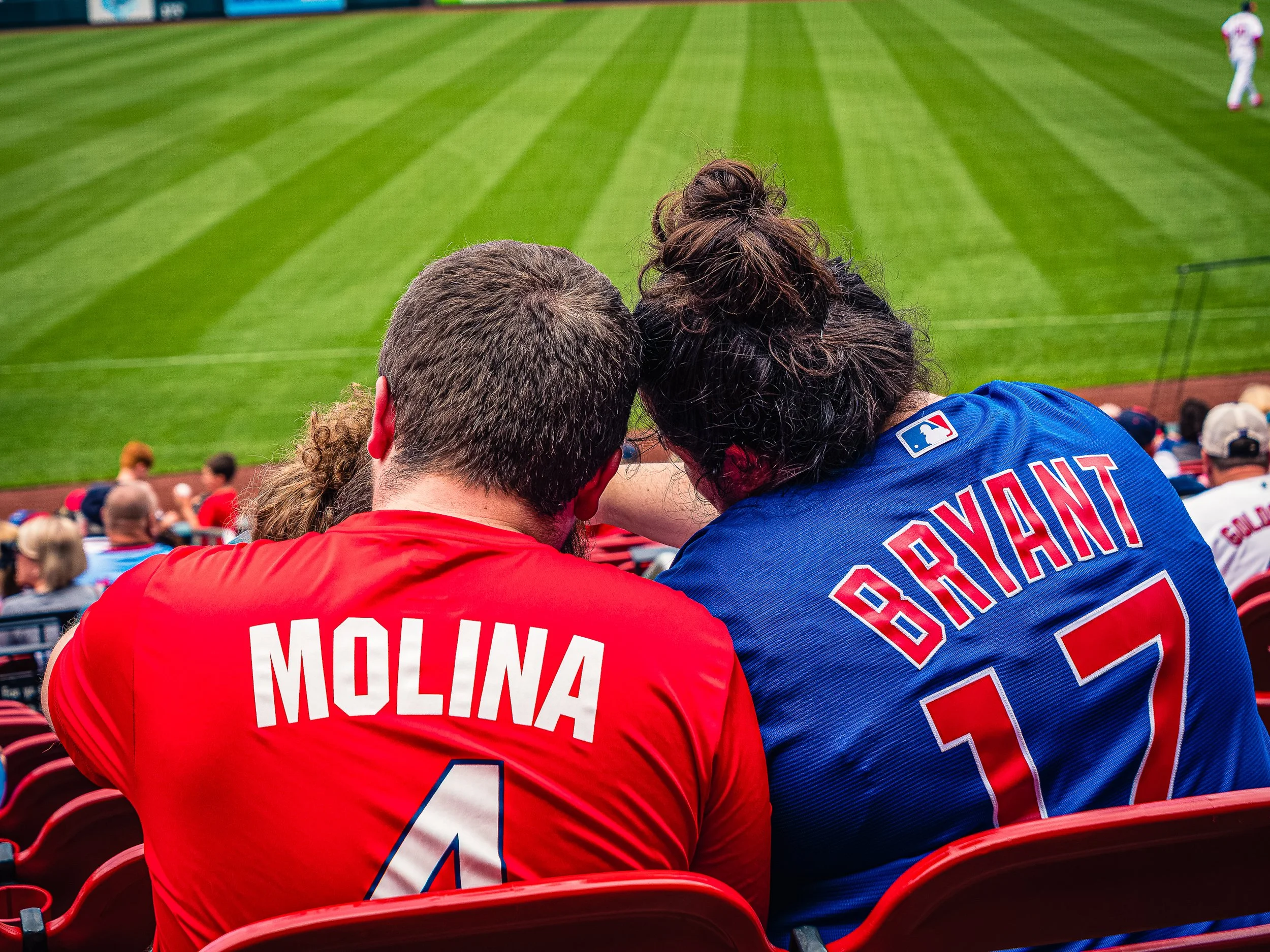 St. Louis Cardinals fan wearing a red Molina number 4 jersey leaning together with a Chicago Cubs fan wearing a blue Bryant number 17 jersey while watching a baseball game.
