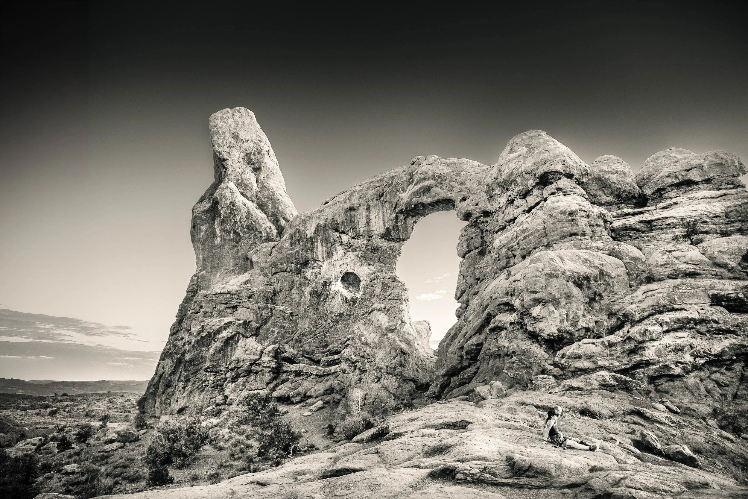 Black and white photograph of a couple seated beneath Turret Arch in Arches National Park, watching the sunset as the sandstone formation towers above them
