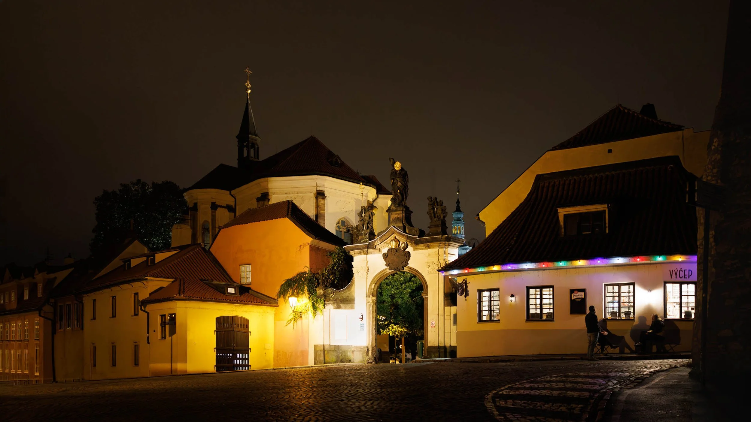Three people gathered outside a warmly lit Prague pub at night beneath historic architecture and an illuminated baroque gateway.