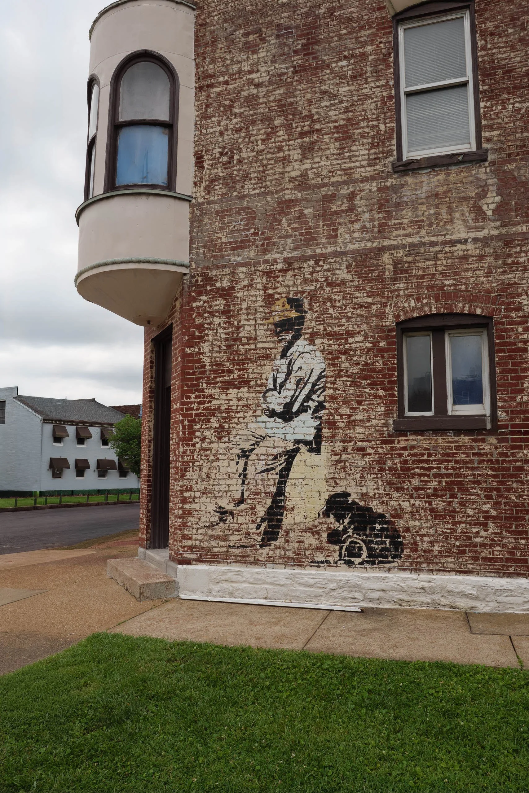 A faded painted mural of a hunter with a shotgun and dog on a weathered brick building corner, with an Art Deco bay window turret above and ghost-sign lettering fading into the brick.