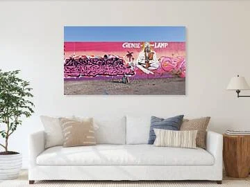 Long magenta-to-peach mural wall in St. Louis with pink graffiti lettering at left, silhouetted palm trees, a seated white-robed figure under the words Genie of the Lamp and a brown lamp at right