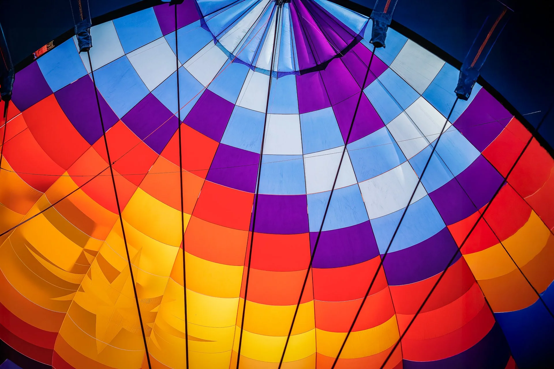 Upward view inside a hot air balloon with curved blue, white, violet, red, orange, and yellow fabric panels crossed by dark rigging cords.
