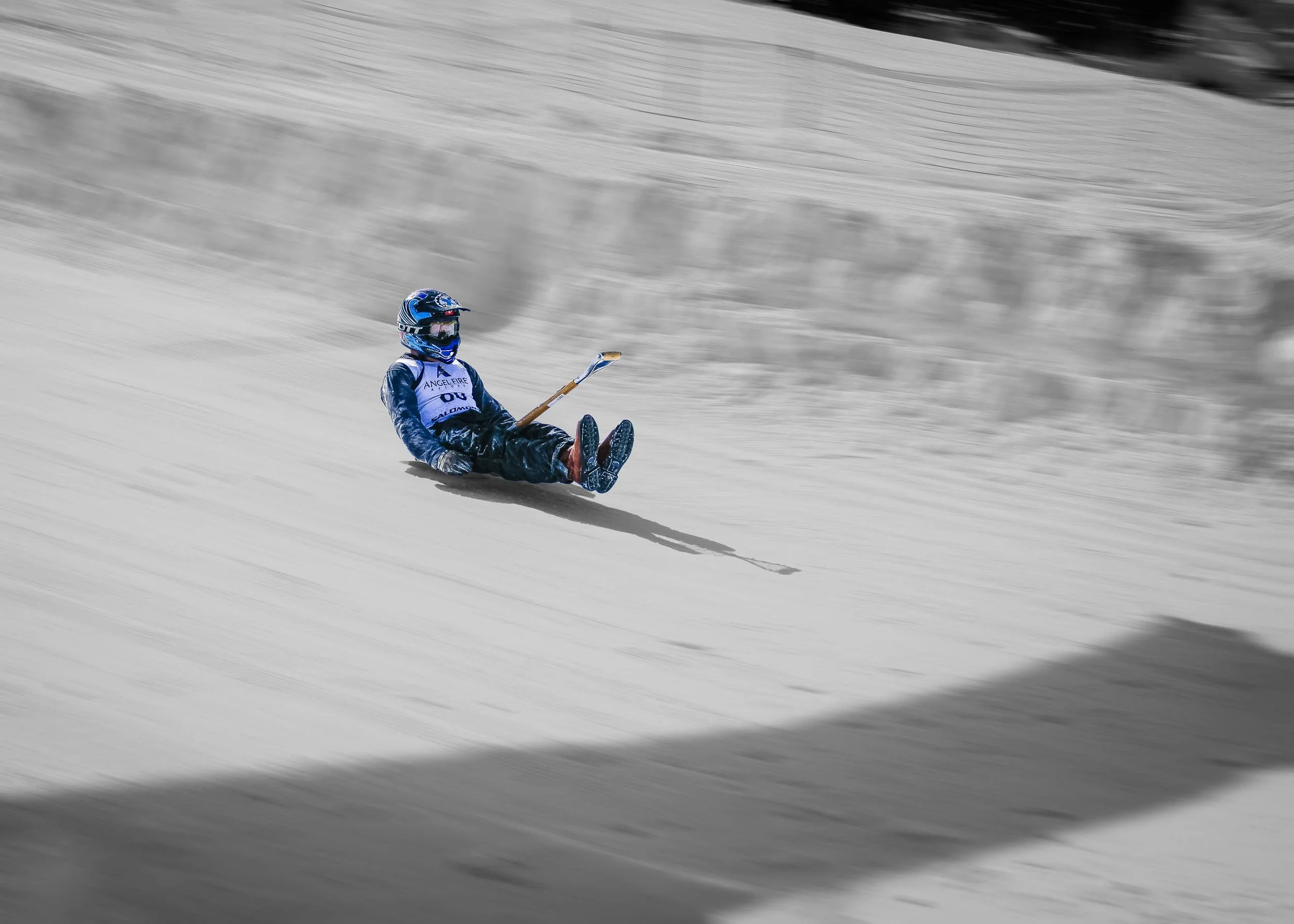 Selective color photograph of a shovel racer in blue gear sliding down a snowy slope after a crash at Angel Fire Resort, New Mexico