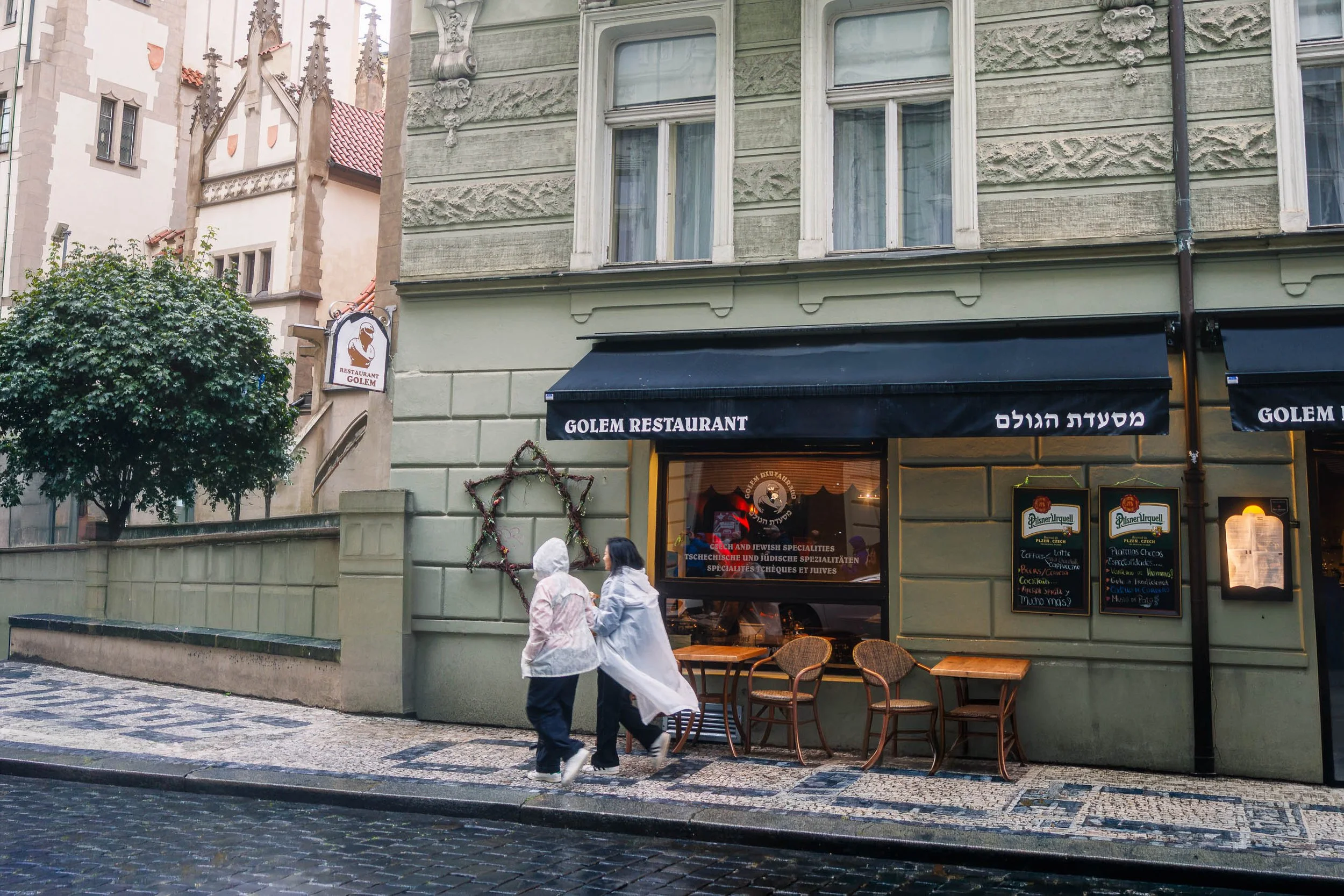 Two figures in rain ponchos walk past the Golem Restaurant and a Star of David in Prague's historic Jewish Quarter on a rainy day.