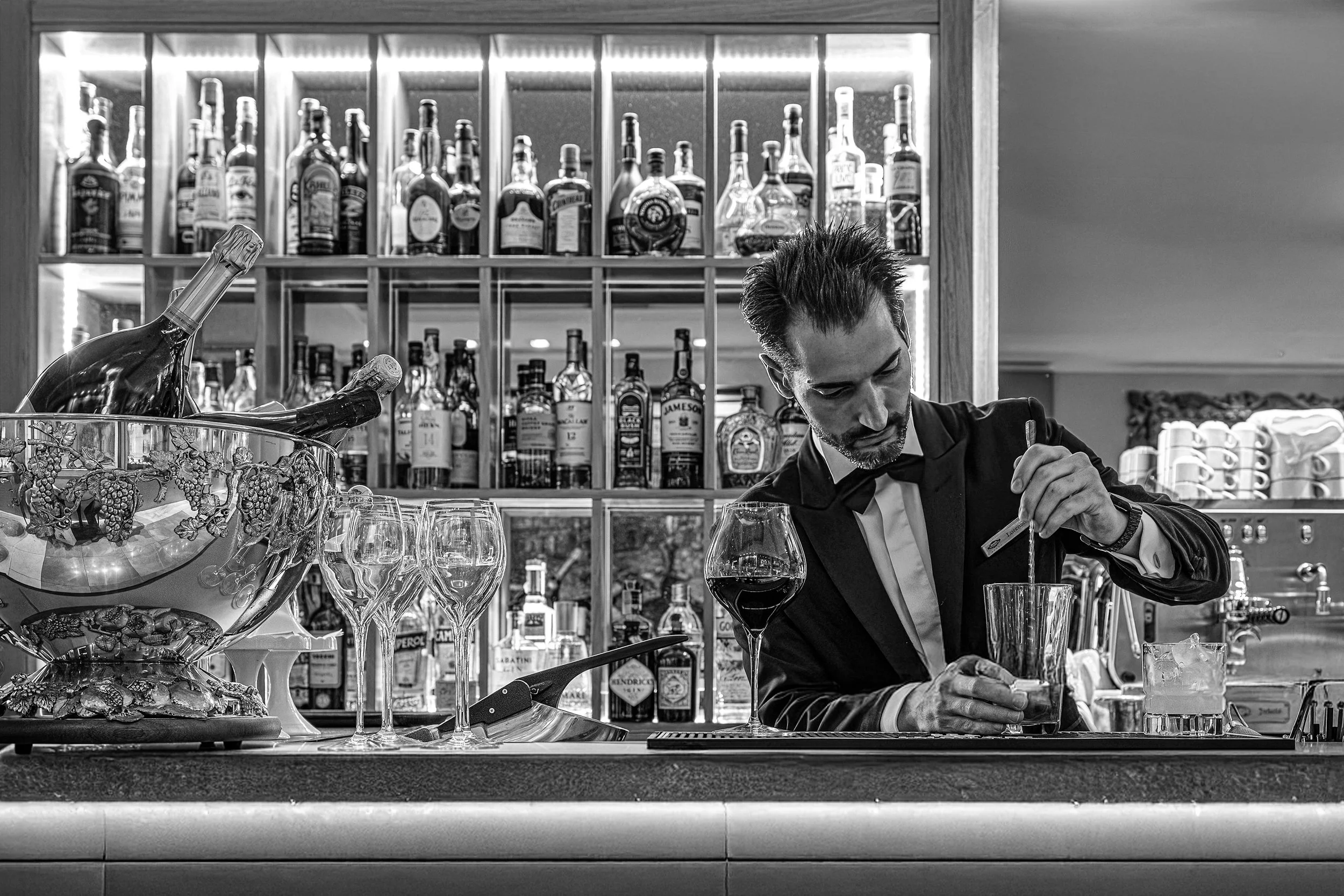 A bartender preparing a drink behind a bar with liquor bottles on shelves in the background. The image is in black and white.