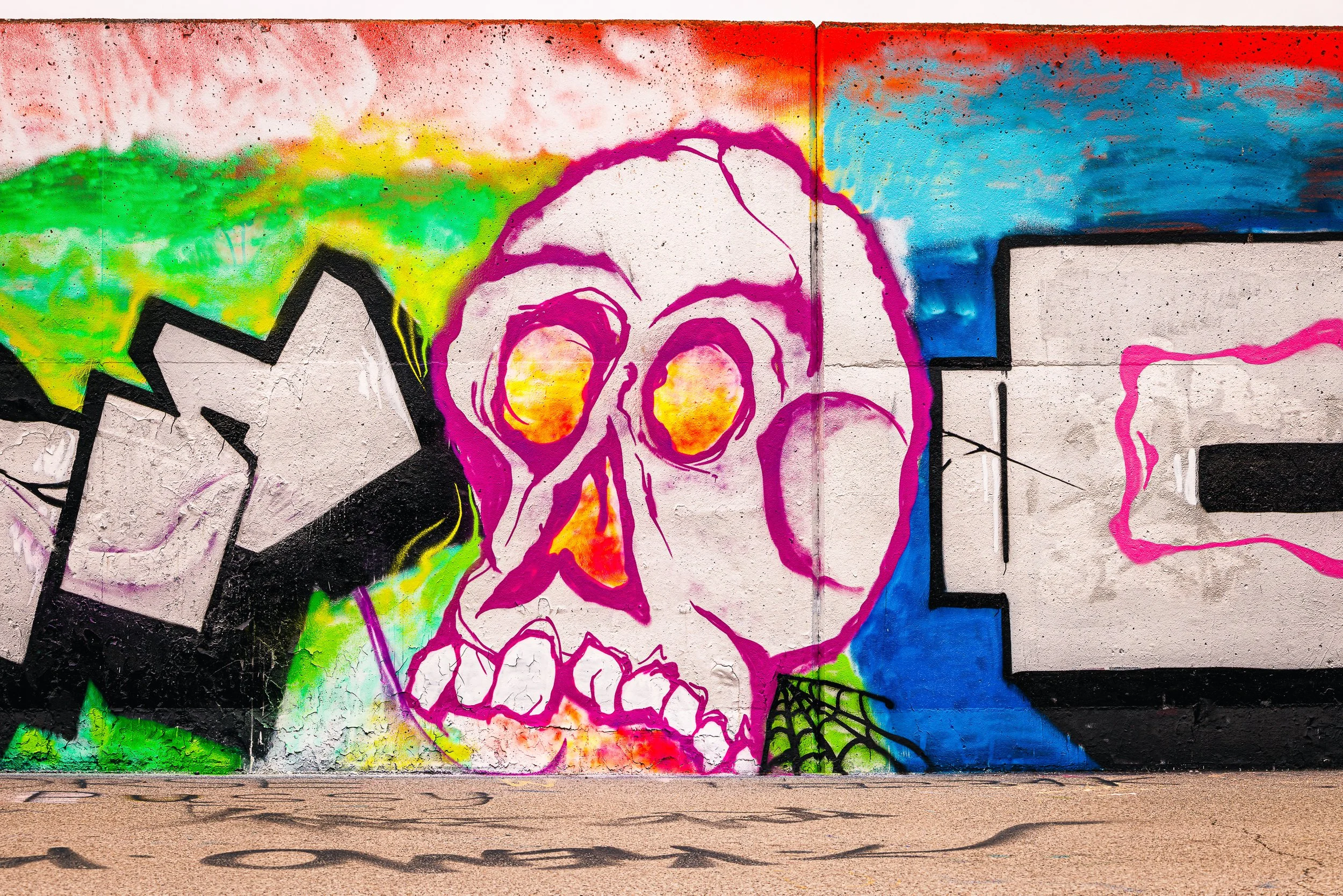 Colorful graffiti mural featuring a pink and orange skull with hollow eyes, outlined in purple, set against a vibrant background of green, yellow, blue, red, and black spray paint on a concrete wall.