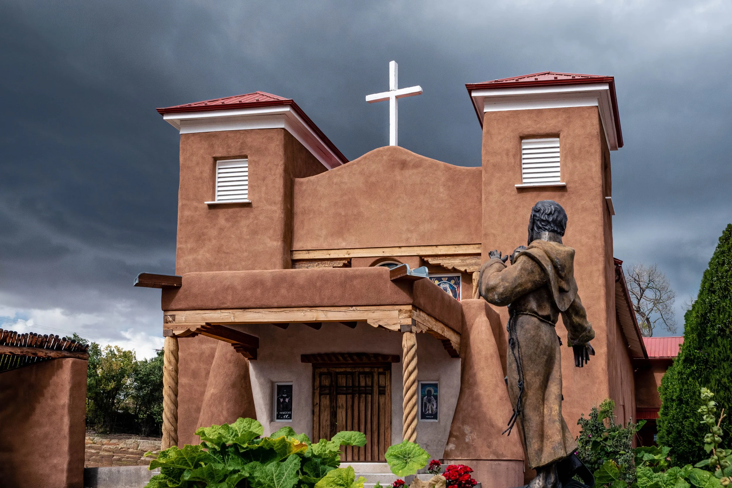 Adobe church and bronze statue beneath storm clouds in Peñasco, New Mexico.
