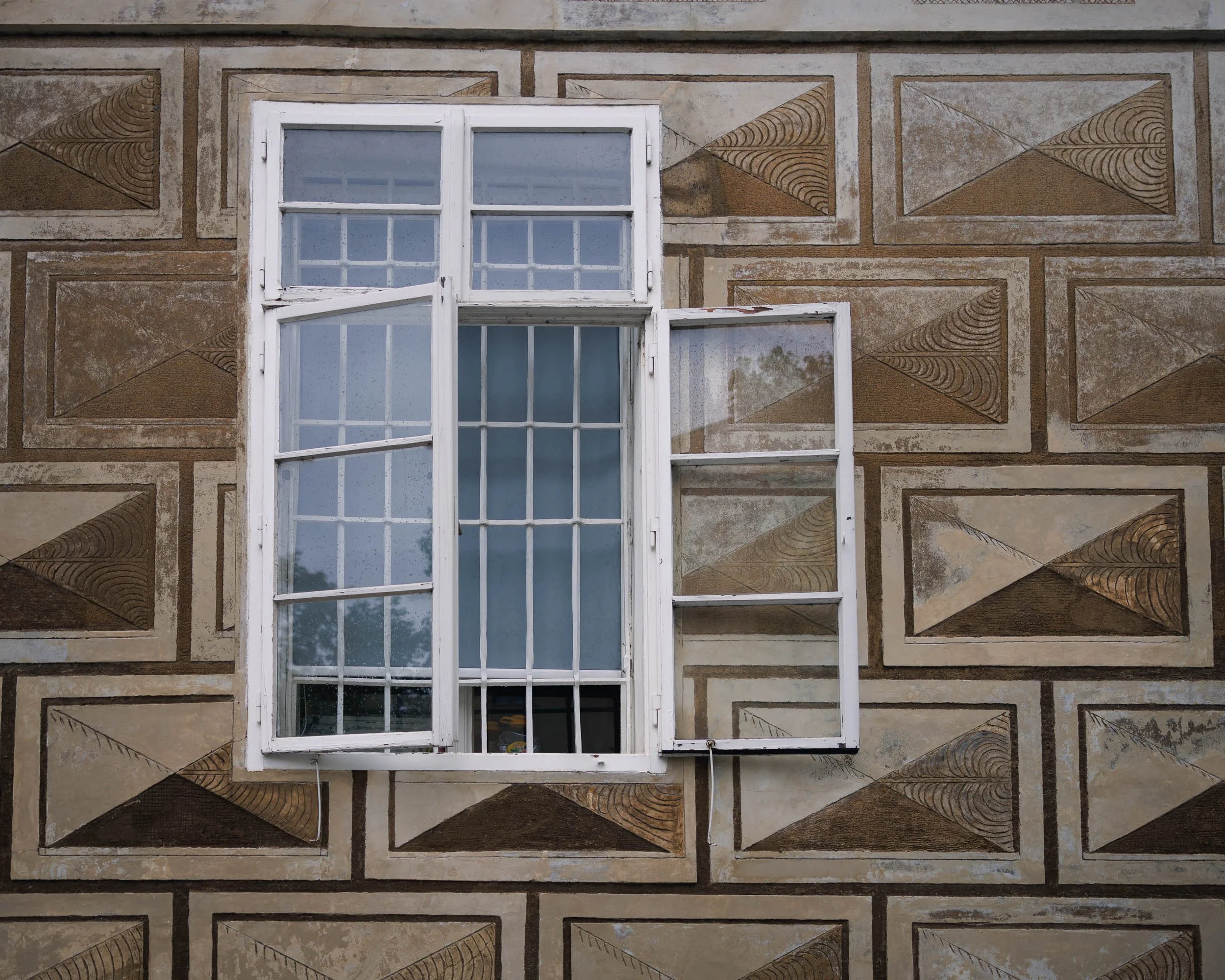 Fine art photograph of an open window interrupting a geometric historic façade in Prague, exploring pattern, architecture, and quiet human presence.