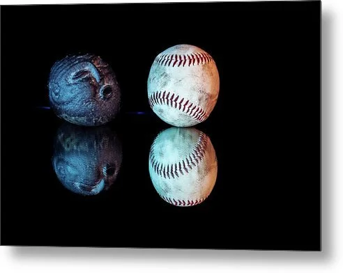 Color still-life photograph of a carved stone face appearing to blow toward a worn baseball, illuminated with blue and amber light and reflected against a black background.