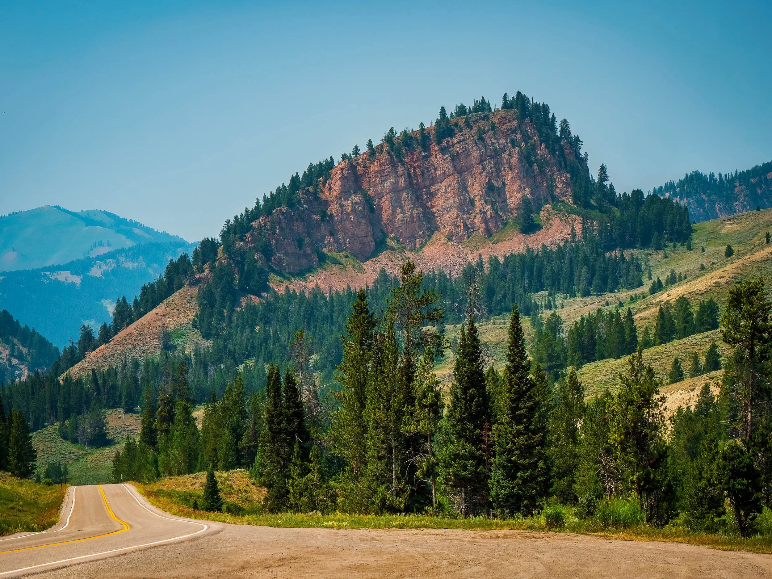 A two-lane mountain highway curves toward a sunlit red sandstone cliff rising above pine forest, photographed on the road into Yellowstone National Park.