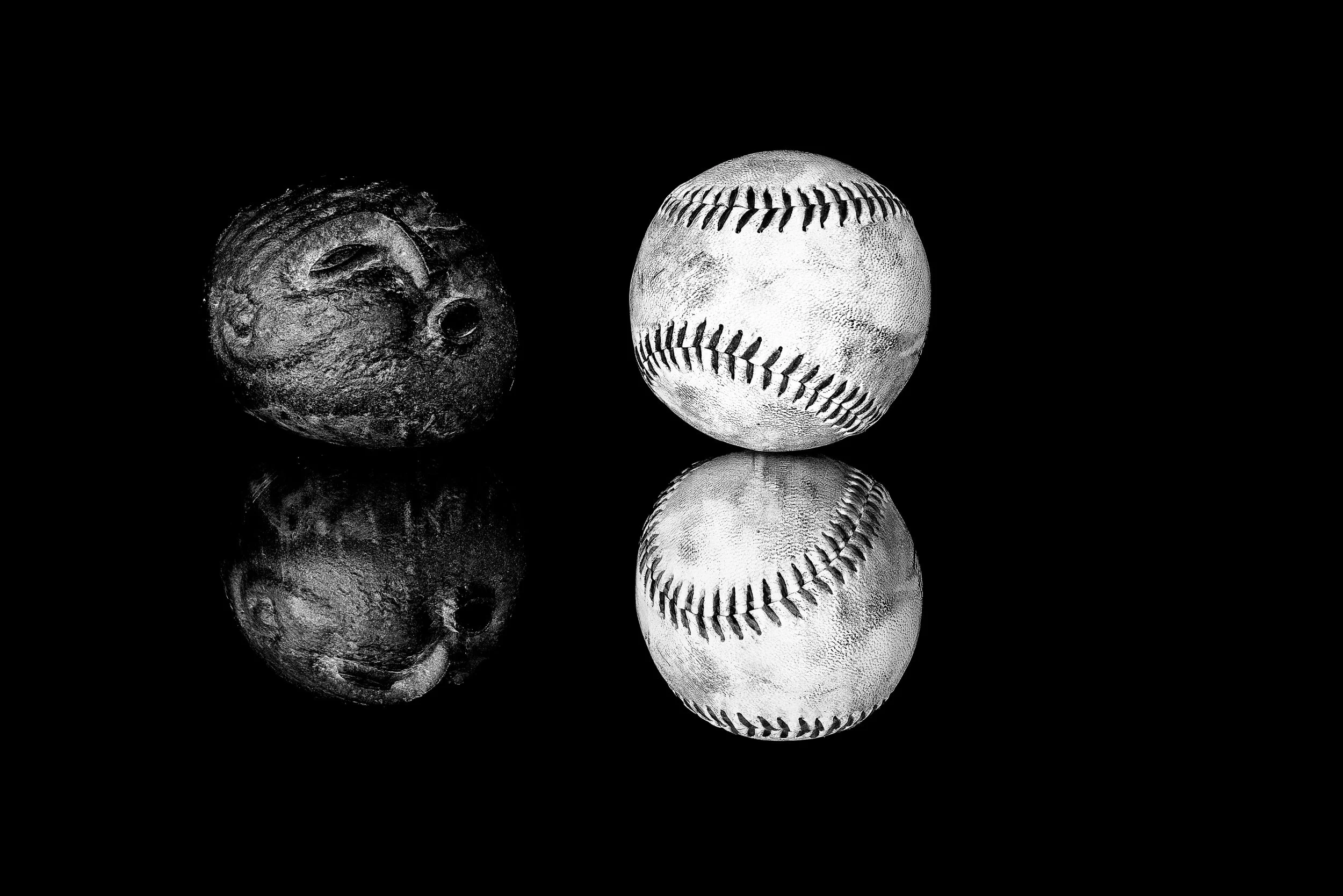 Black-and-white still life of a carved stone face appearing to blow toward a worn baseball, with both objects reflected against a deep black background.