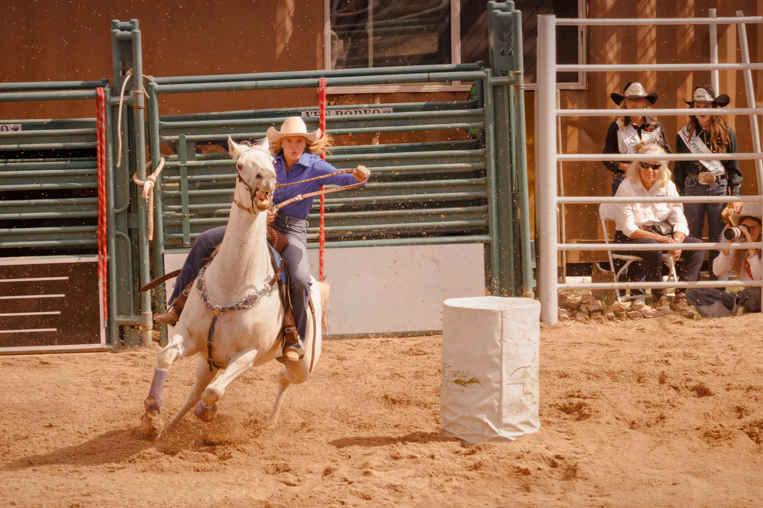 A woman barrel racer on a white horse at full gallop rounds a barrel at the Evergreen Rodeo in Colorado, blue shirt vivid against the arena panels, hair and arena dust flying.
