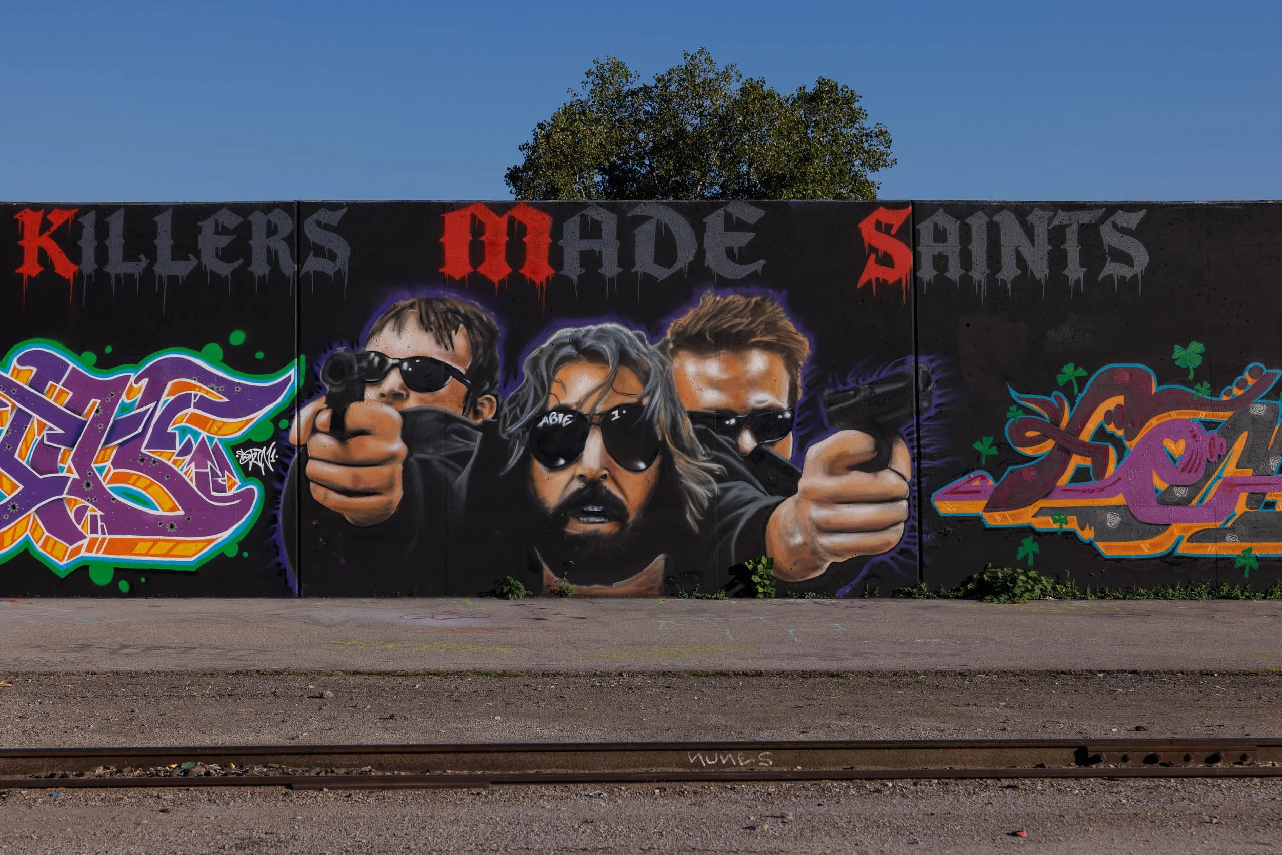 Mural of three figures in dark sunglasses with arms extended toward the viewer on a black wall in St. Louis, red and gray dripping lettering above and purple graffiti letters at left