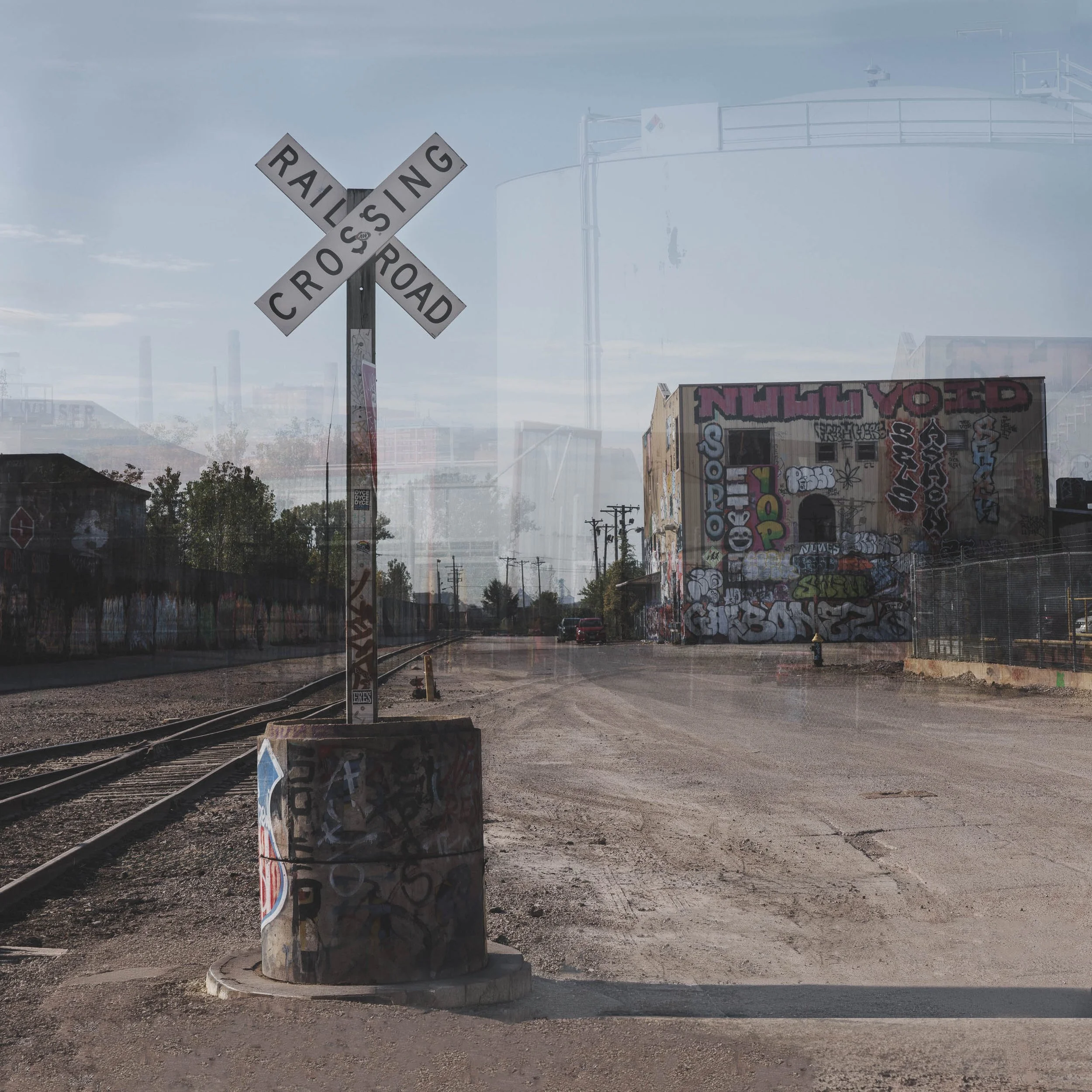 Railroad crossing sign sharp against layered ghost images of a tank and graffiti warehouse, St. Louis, in blue and tan