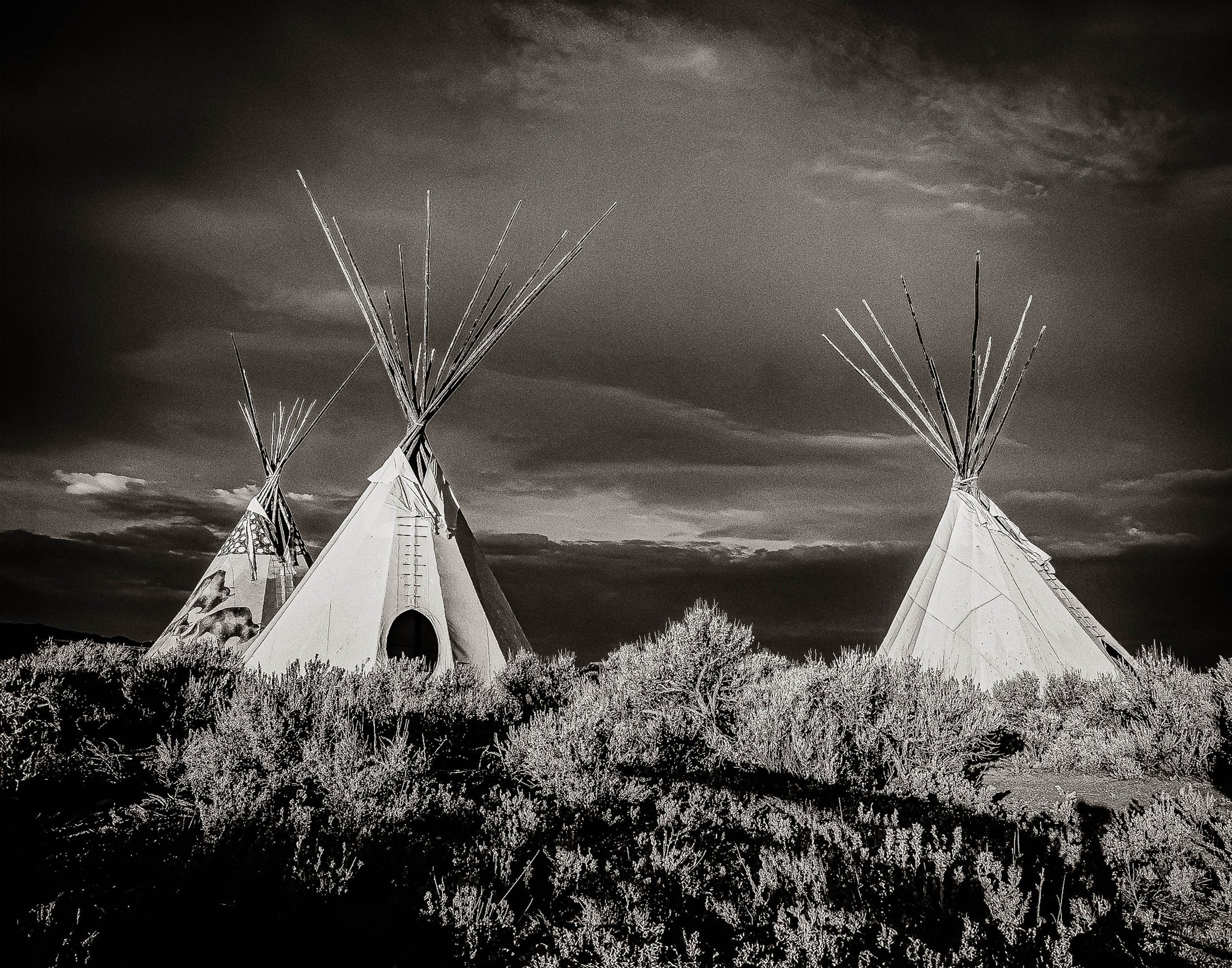 Black and white photograph of three tipis beneath dramatic storm clouds in the American West.