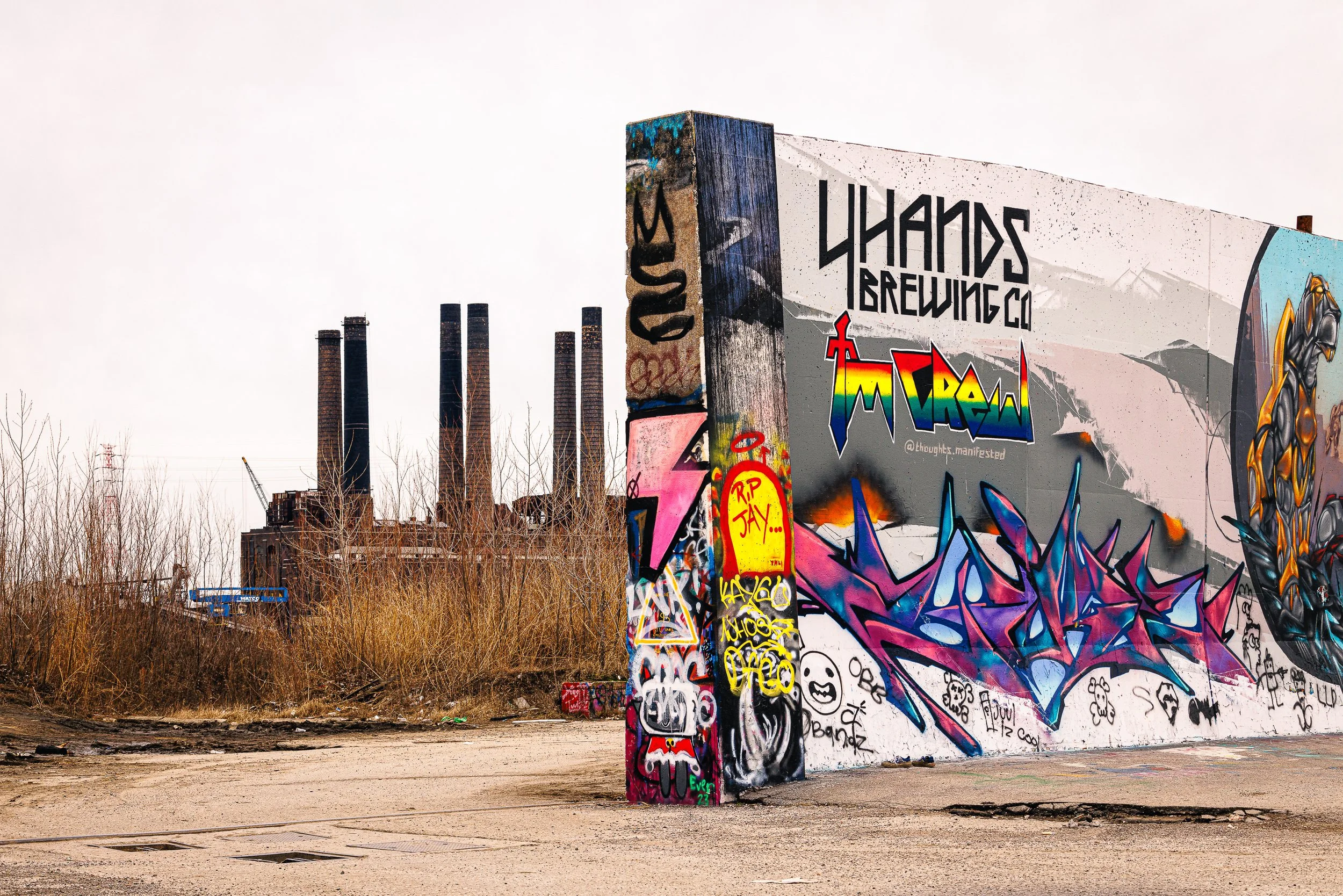 Colorful graffiti art on a large wall and pillar in an urban setting with industrial buildings and smokestacks in the background, and leafless trees in the foreground.