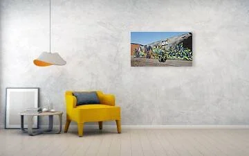 Two riders on a green motorcycle pass a long gray mural wall with gold, blue and violet-green lettering, St. Louis shown in room with chair