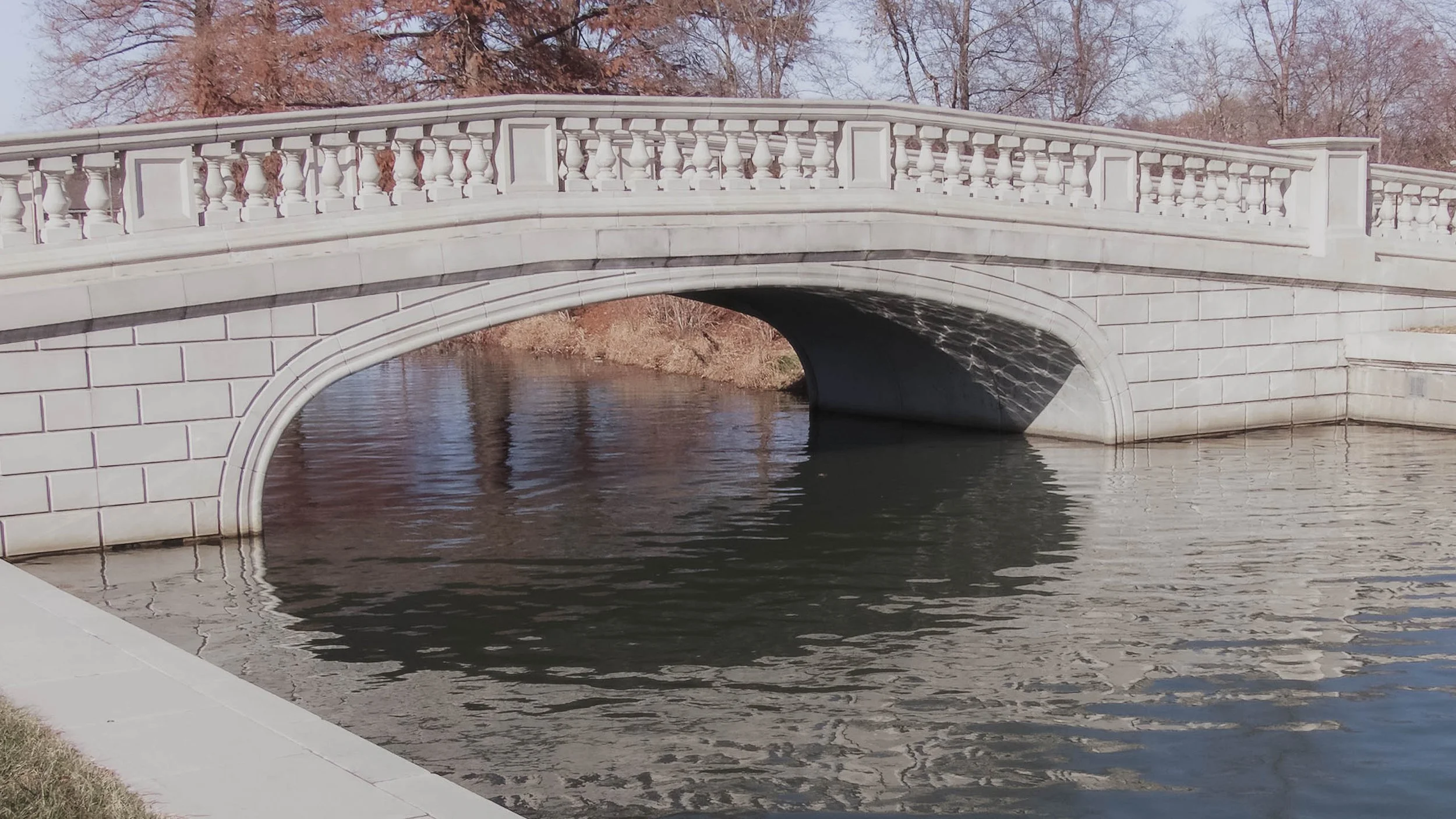 Architectural photograph of a pale stone bridge reflected in calm water beneath soft winter light.