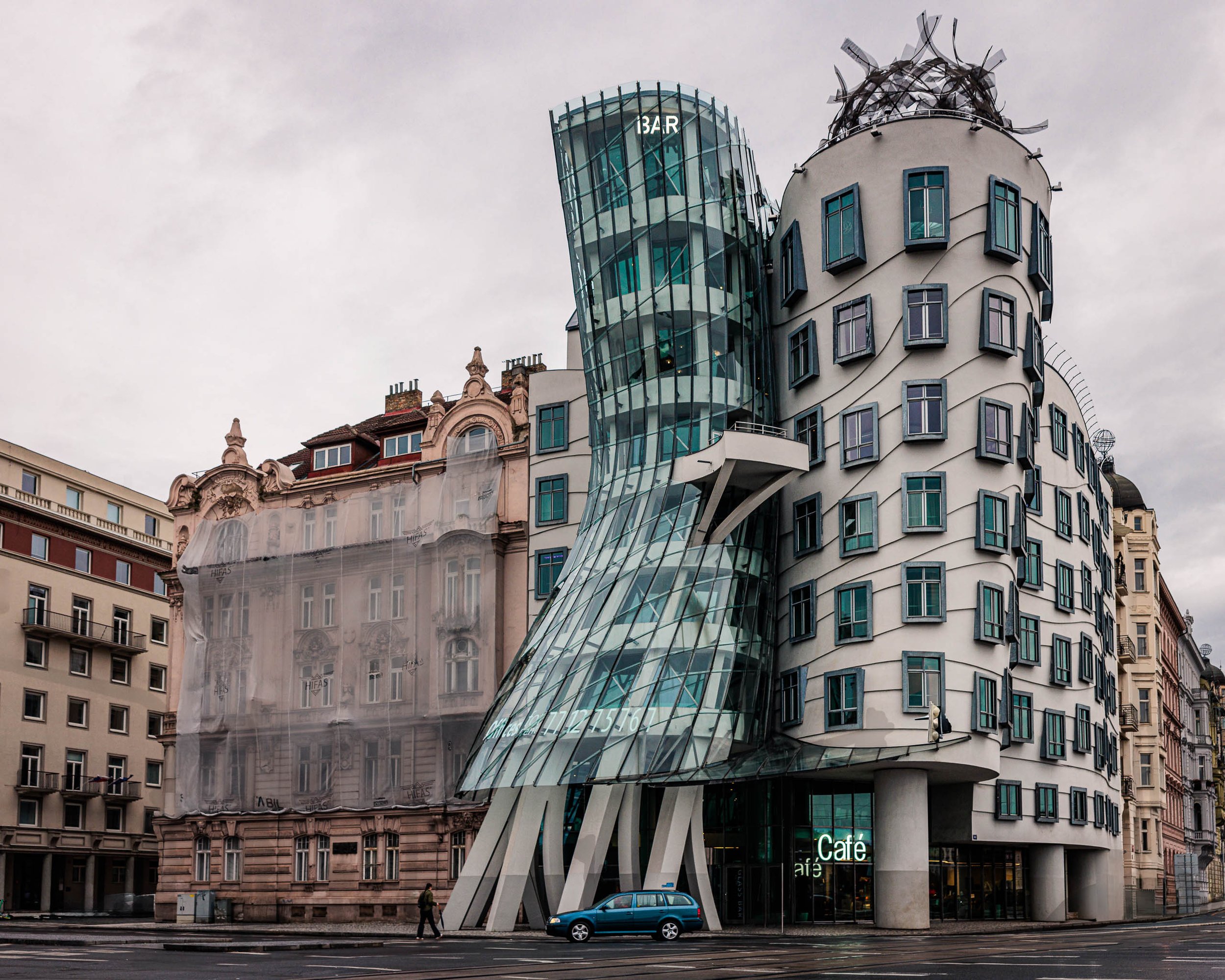 The Dancing House in Prague stands beside a historic nineteenth-century building under an overcast sky, highlighting the contrast between modern and historic architecture.