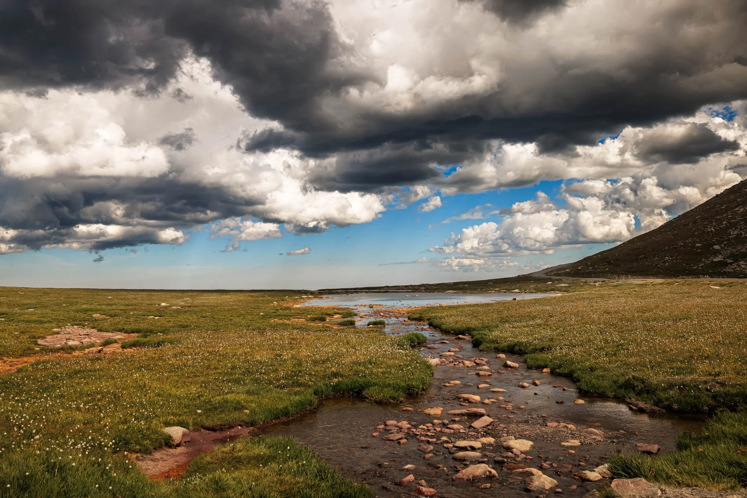 Dark storm clouds building over alpine tundra, a rust-stained mountain stream leading to a lake, and a thin highway on the slope at right, near Mount Blue Sky, Colorado