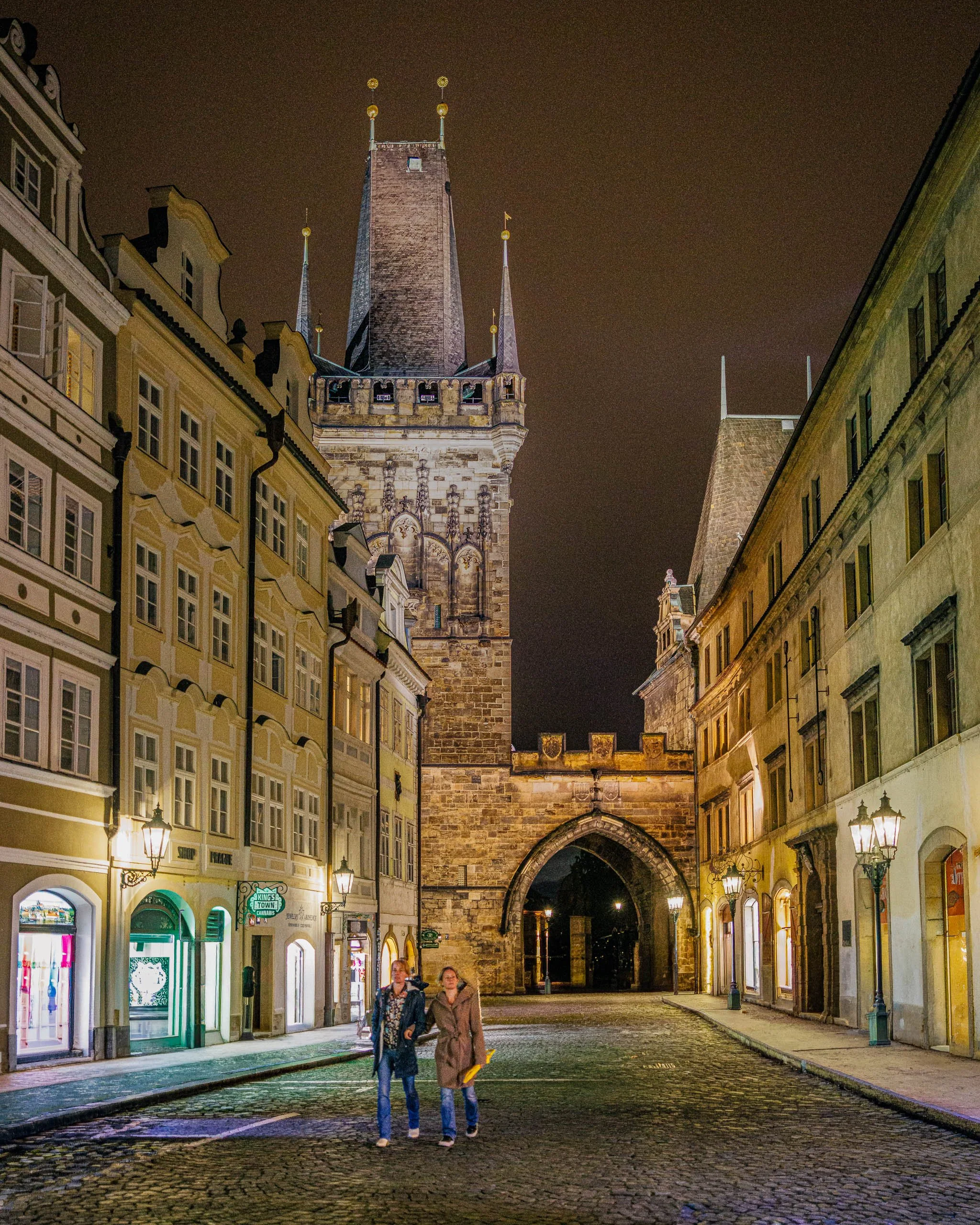 Two pedestrians walk beneath Prague's illuminated Old Town Bridge Tower on a quiet night street lined with historic buildings and warm street lamps.