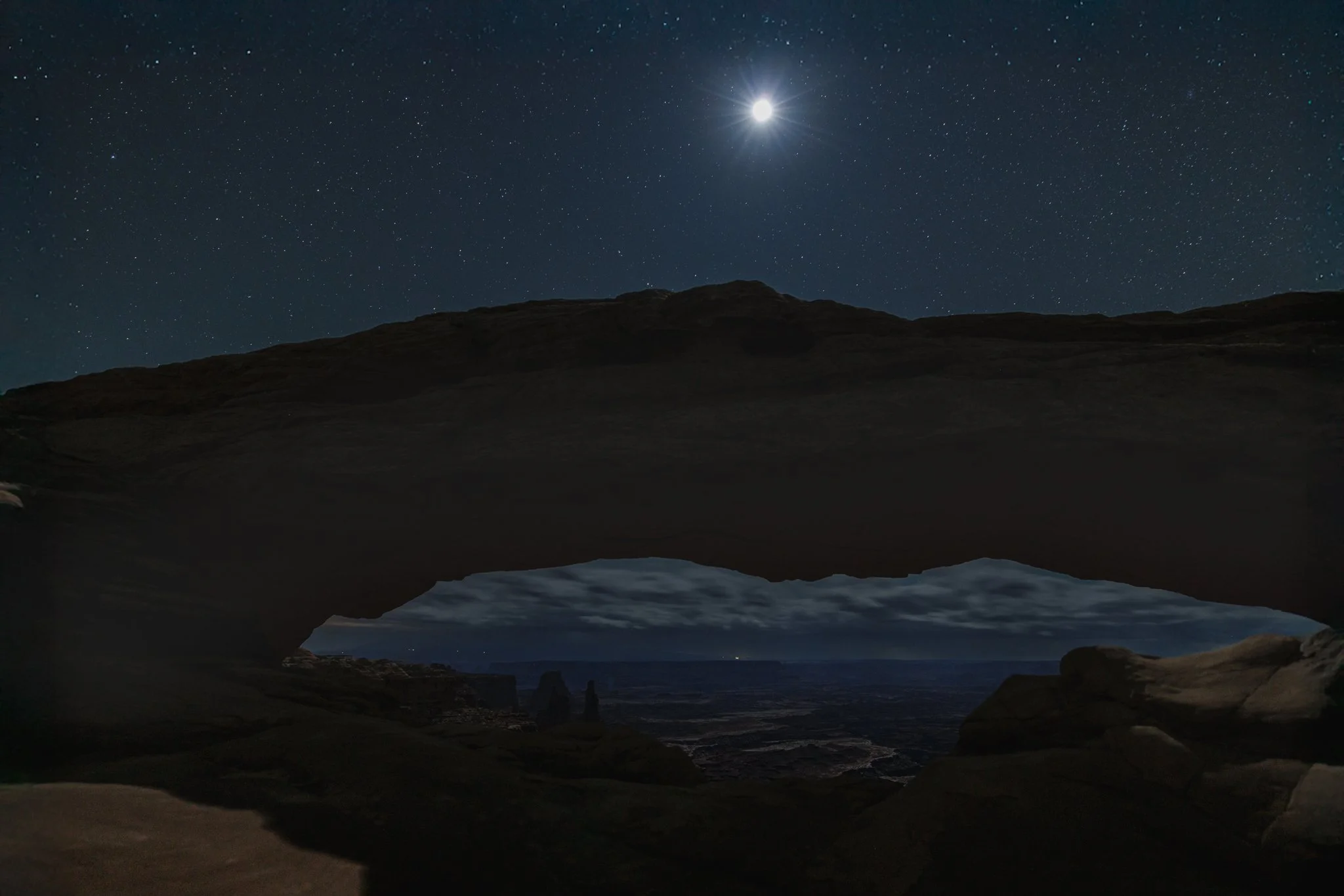 Fine art landscape photography print capturing Mesa Arch under a clear midnight sky filled with stars, with a small still puddle of water reflecting the light on the right.