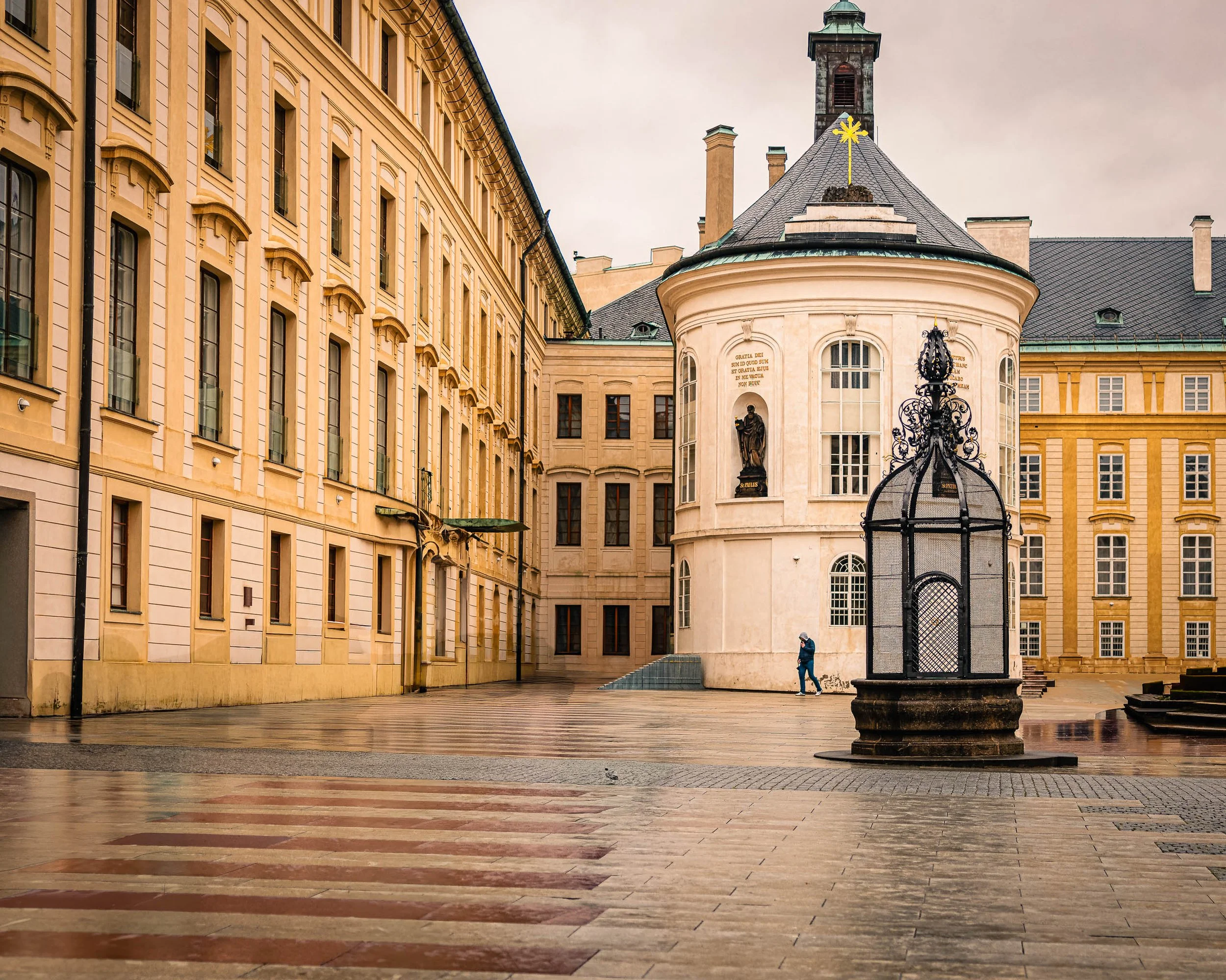 A lone figure walks across a rain-soaked courtyard at Prague Castle surrounded by historic architecture.