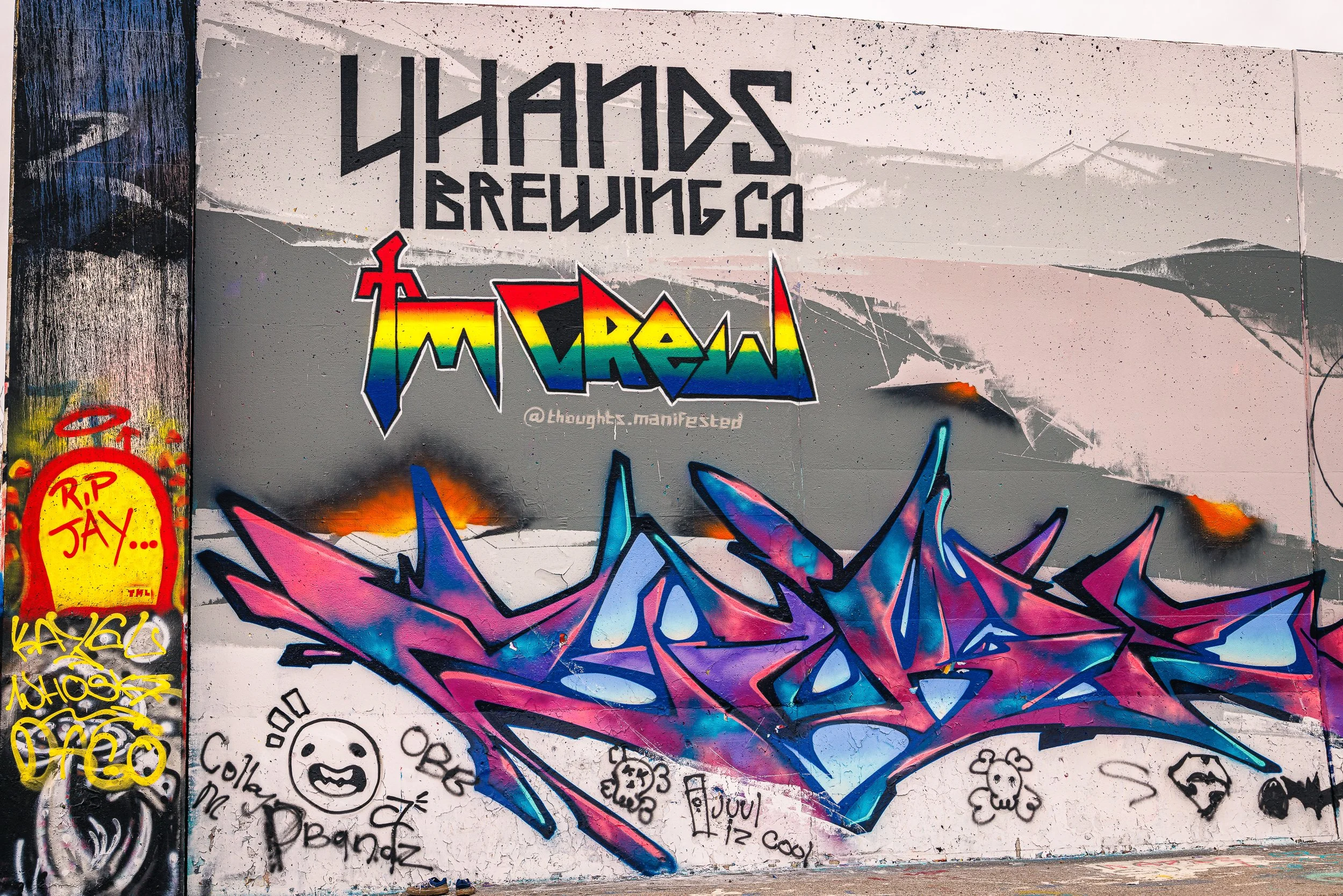 Graffiti mural on a wall with a large, colorful, stylized paint splatter at the center. The top part of the wall features the text 'THANKS BREWING CO' in black, outlined letters. The lower part contains various smaller graffiti tags and doodles inclu
