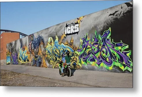 Two riders on a green motorcycle pass a long gray mural wall with gold, blue and violet-green lettering, St. Louis Shown full size in Metal