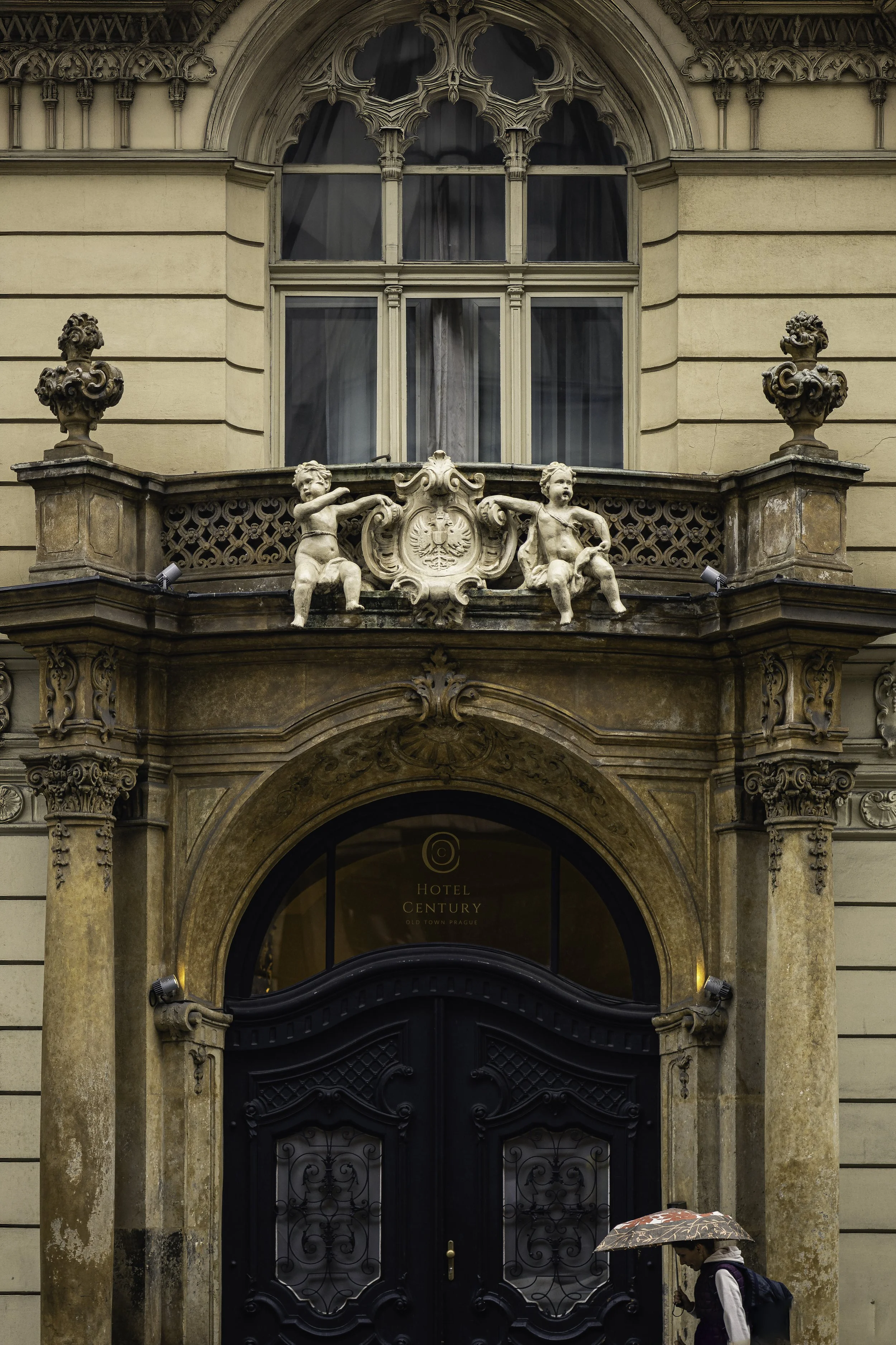 Pedestrian carrying an umbrella past an ornate historic doorway in Prague framed by Baroque stone architecture.