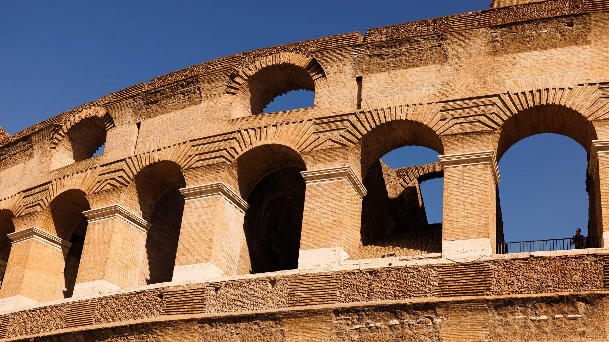Golden-hour photograph of the Colosseum's upper arcade in Rome, Italy, with sunlit stone arches, deep shadow, and a lone figure for scale.