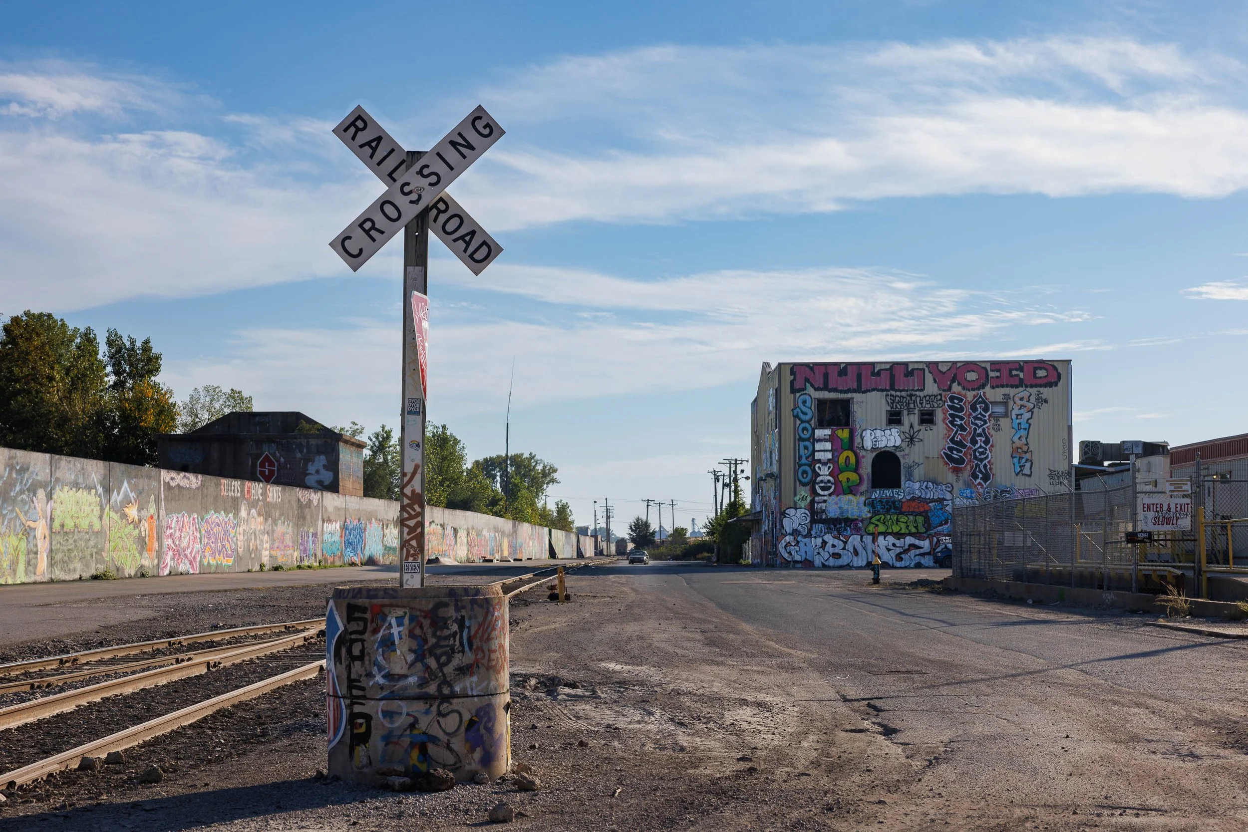 Railroad crossing sign beside converging tracks and an empty industrial street, with graffiti walls and blue sky, St. Louis