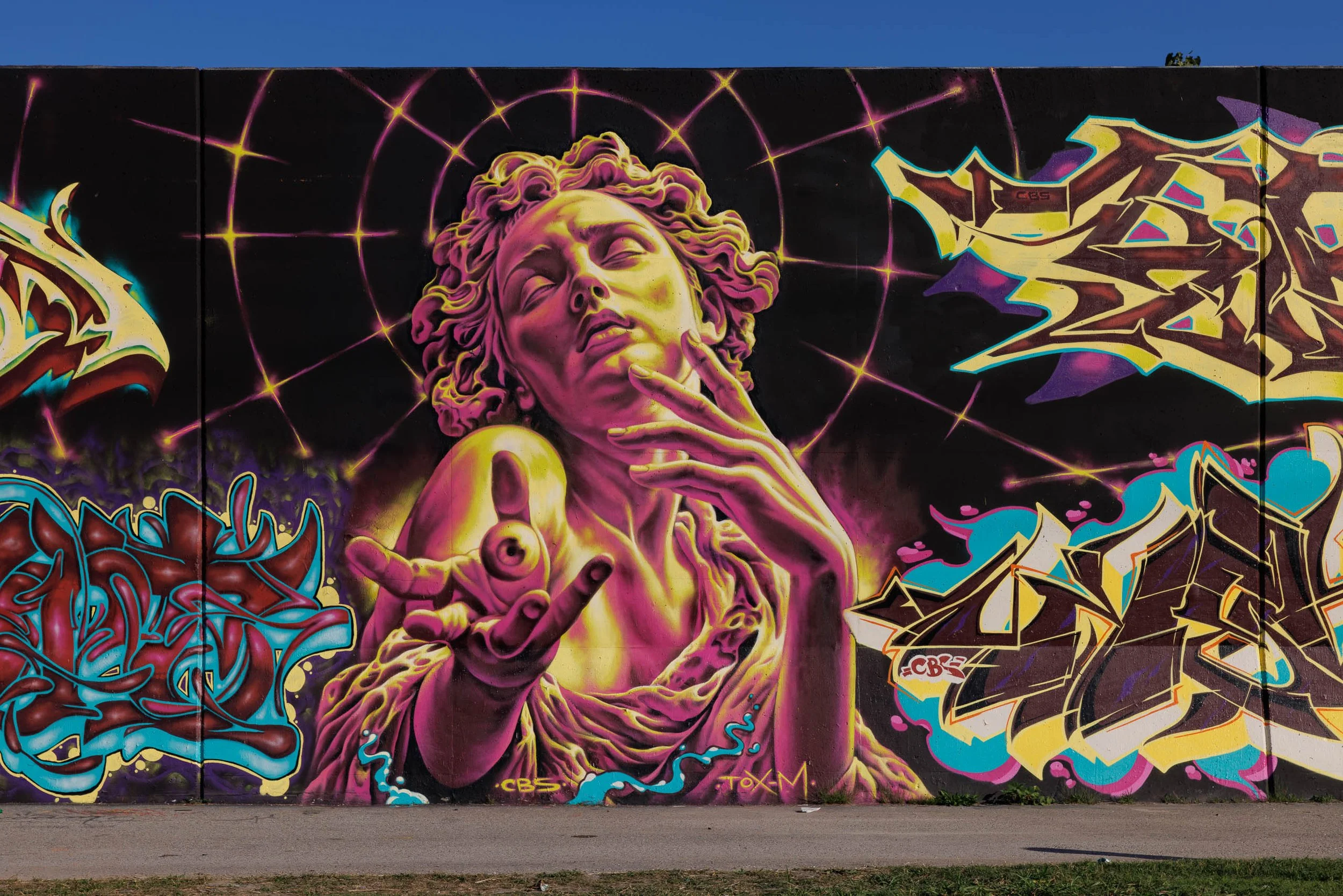 Mural of a classical sculpture-style figure in magenta and golden yellow with curled hair and closed eyes, one hand at the chin and an open palm holding an eyeball.