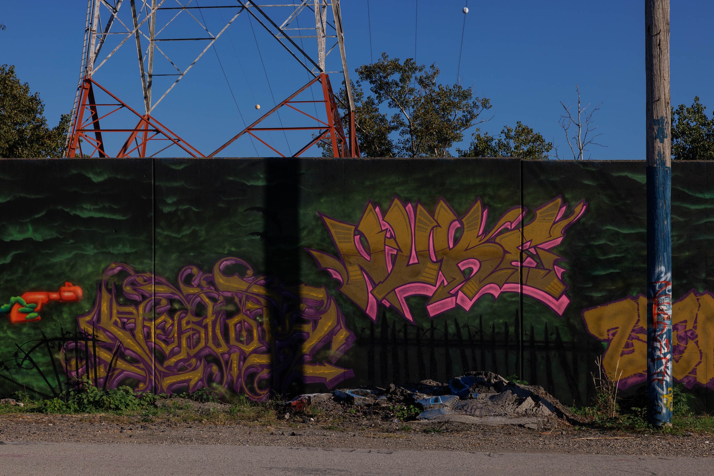 Red and white lattice transmission tower rising above a long deep-green mural wall with gold and pink graffiti lettering and a sticker-covered blue utility pole in St. Louis, under a blue sky