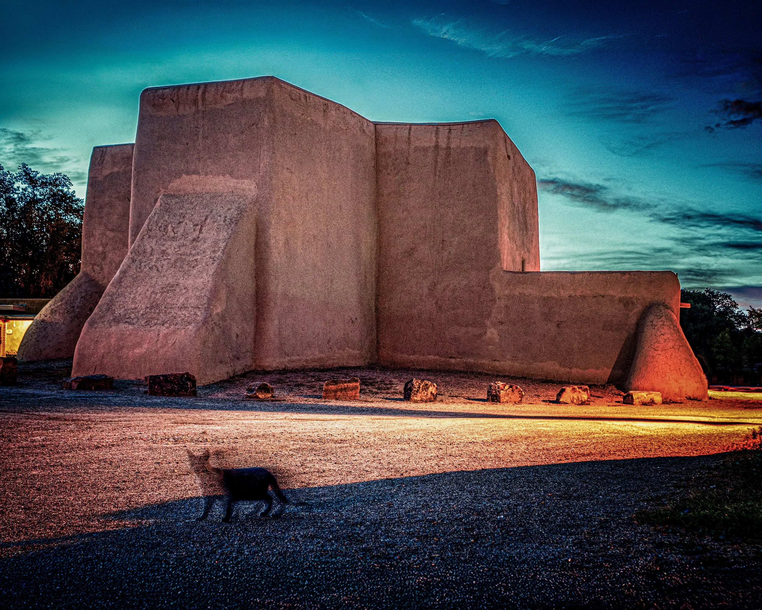 Rear view of the San Francisco de Asís Mission Church in New Mexico at dusk, featuring dramatic shadows on the adobe walls and a clear desert sky.