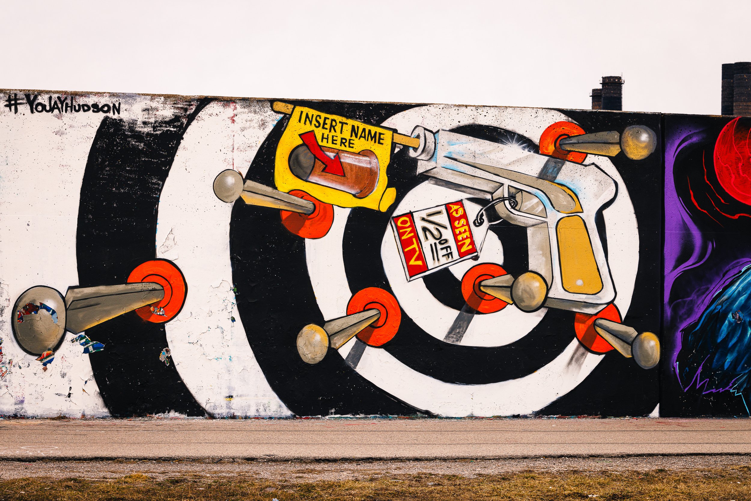 Colorful mural wall featuring abstract shapes, spray paint effects, and a yellow sign with the text 'Insert Name Here.' The mural contains various tools and elements, with a mix of black, white, orange, purple, and yellow colors.