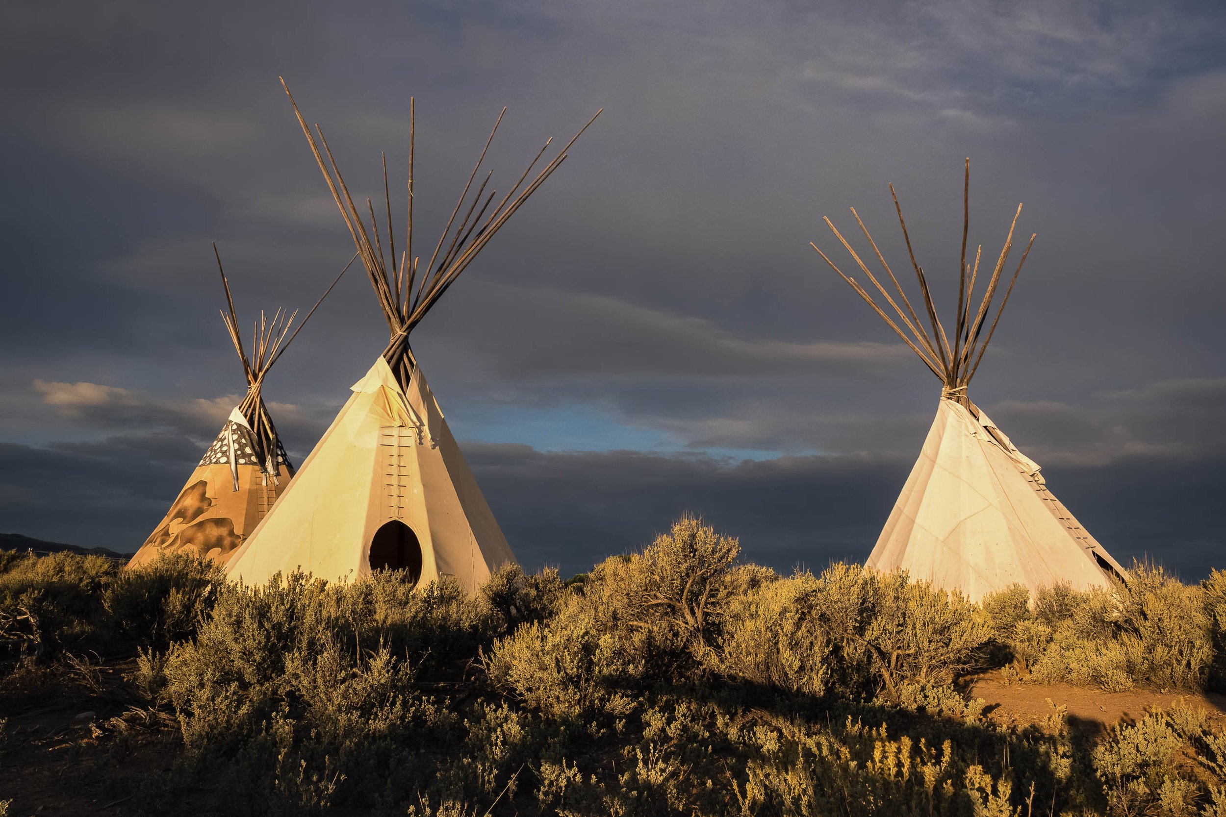 Three tipis illuminated by warm evening light beneath a dark stormy sky, surrounded by sagebrush in a Western landscape.