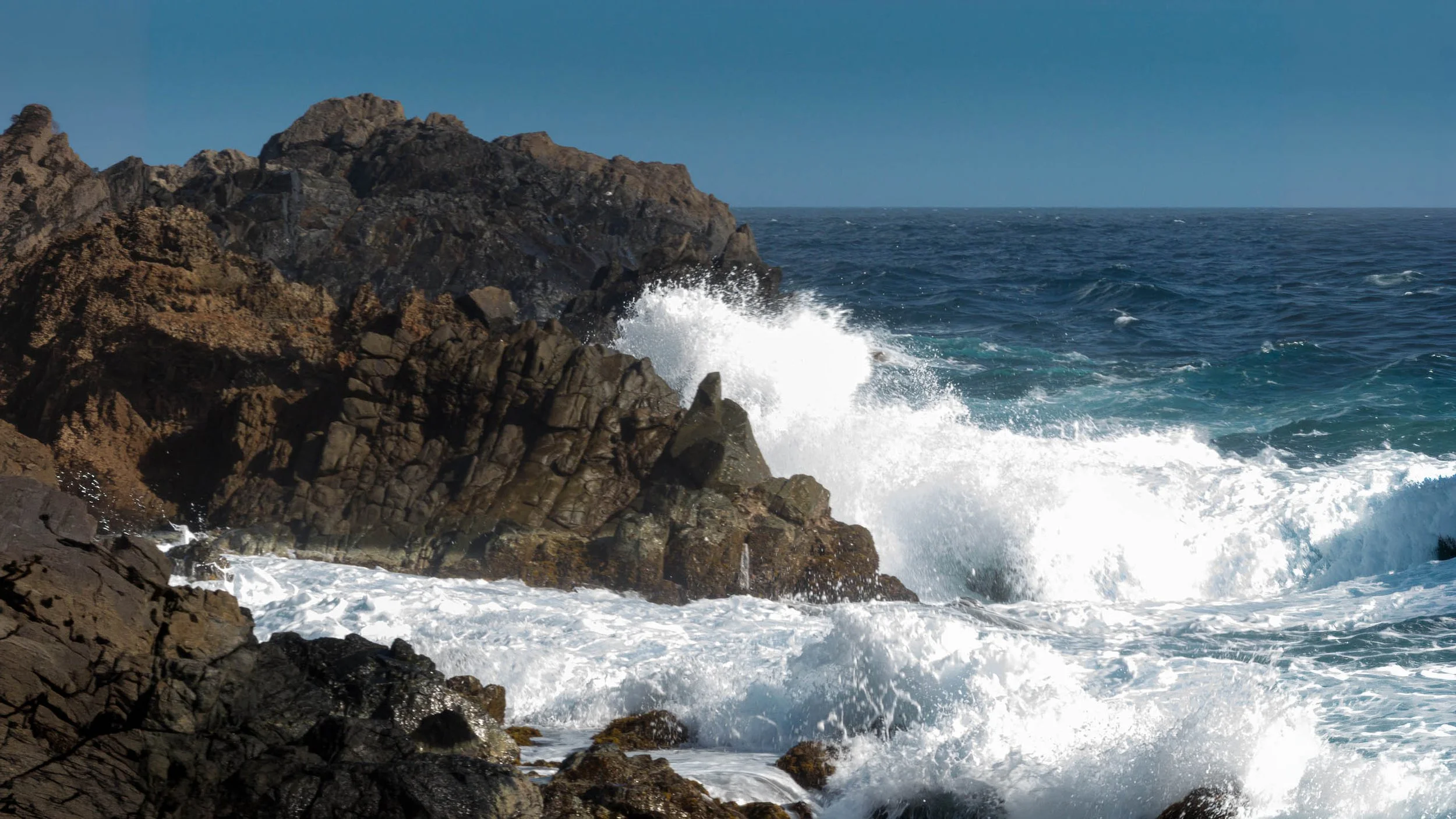 Wave breaking in white spray against dark volcanic rock under a deep blue sky on Aruba's windward coast, navy and white
