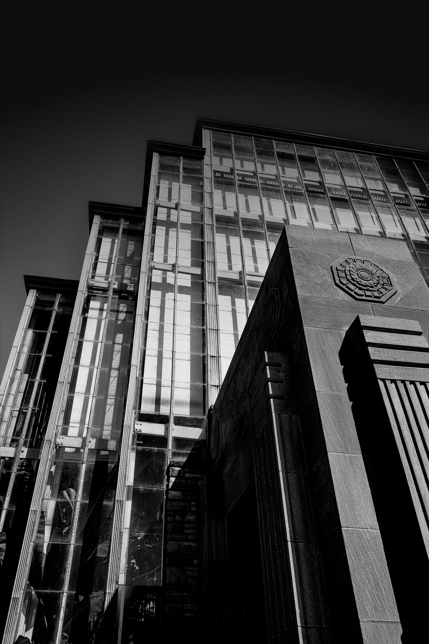 Upward black-and-white view of the Jewel Box in Forest Park, St. Louis, with silver-gray stepped glass panels and a pale carved stone entrance beneath a charcoal sky.