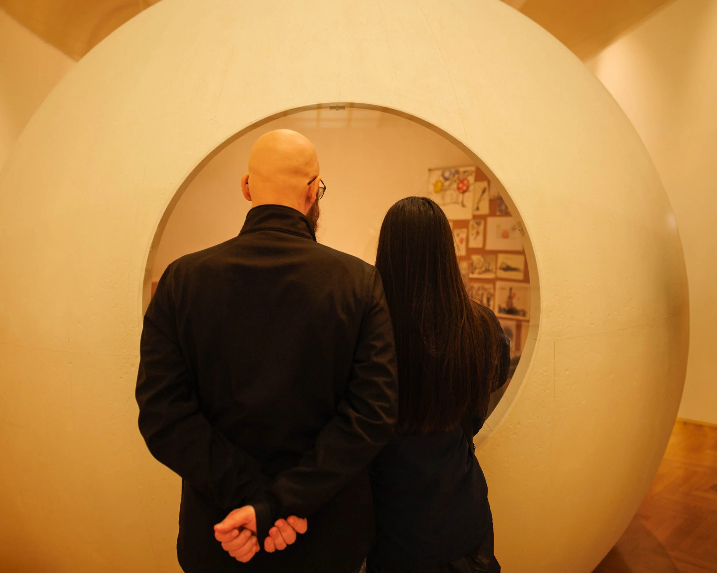 Two visitors stand together inside a Prague gallery, framed by a large circular architectural opening while viewing an exhibition beyond.