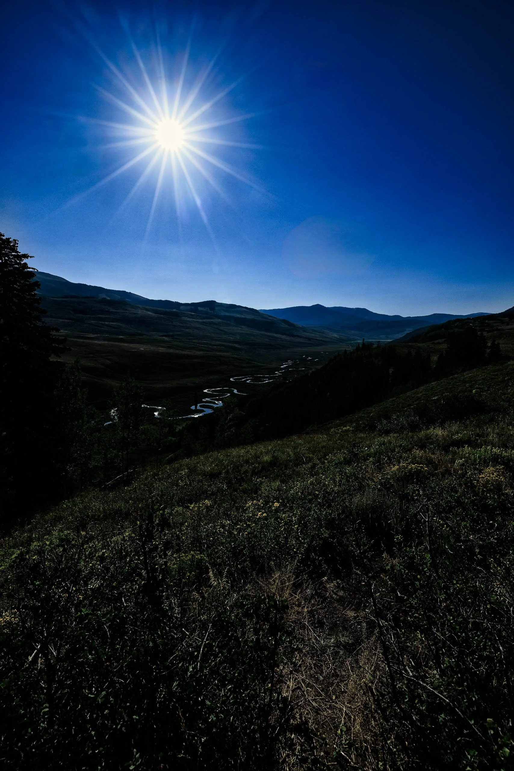 Radiant sunstar over a silhouetted Colorado mountain valley with a winding river below, near Crested Butte