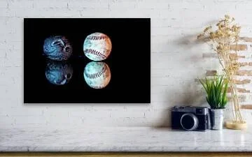 Color still-life photograph of a carved stone face appearing to blow toward a worn baseball, illuminated with blue and amber light and reflected against a black background.