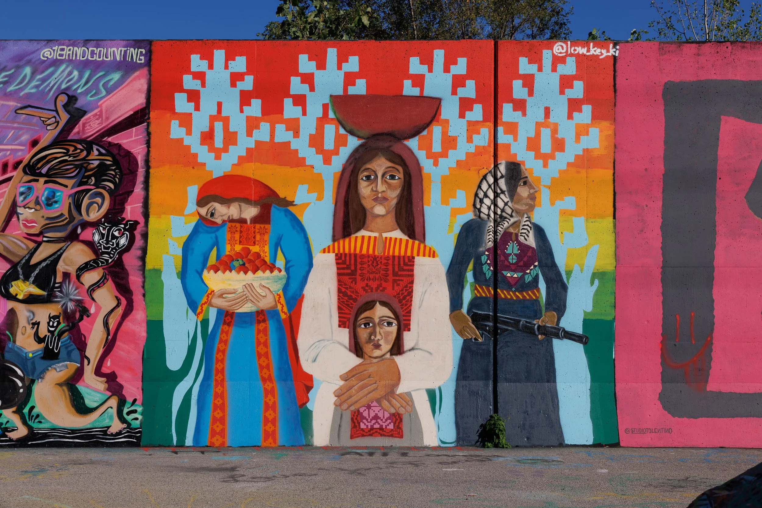 Mural of three standing figures in embroidered dress before horizontal rainbow bands and stepped pale-blue motifs on a wall in St. Louis, the center figure balancing a bowl on her head