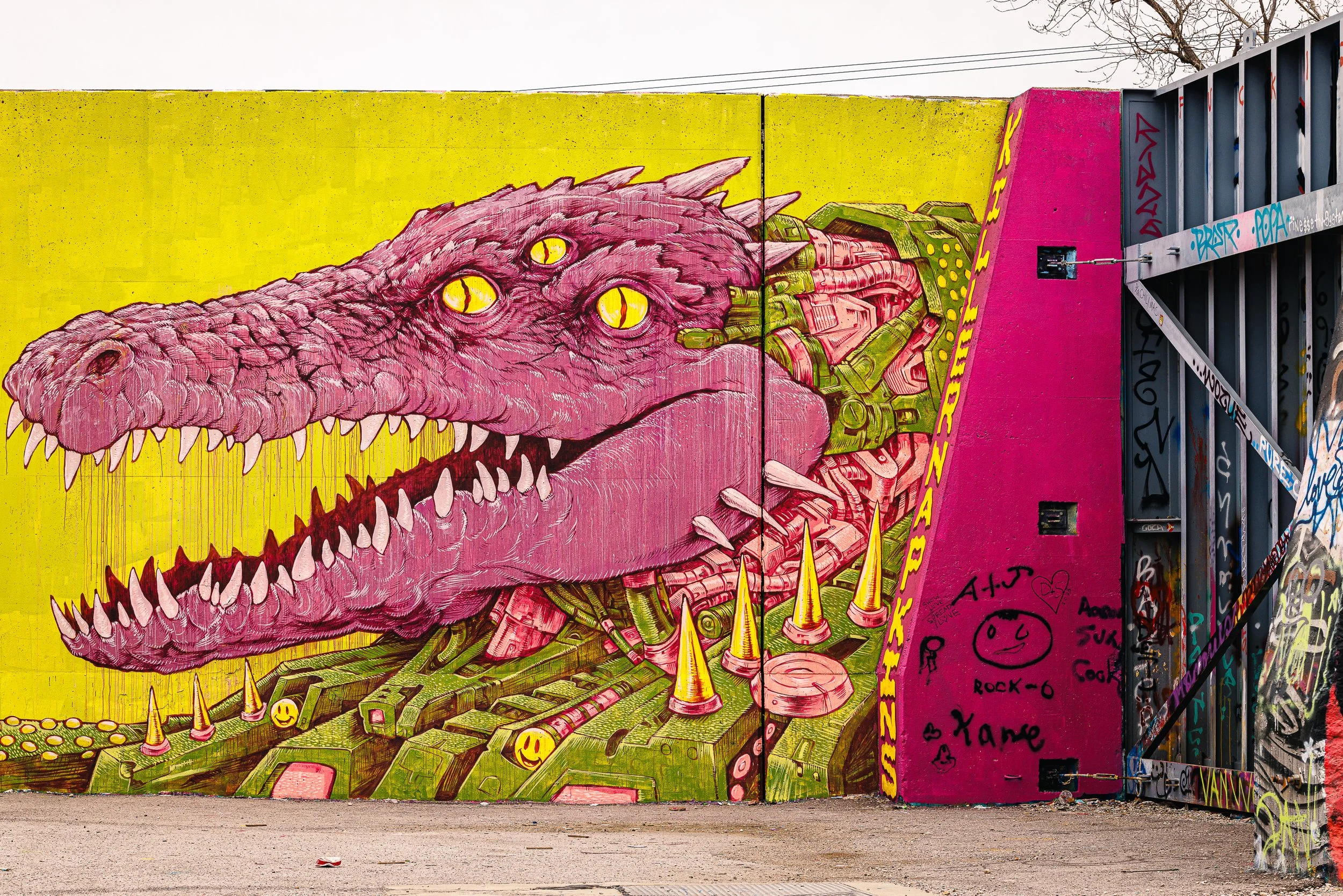 Colorful graffiti of a pink dinosaur with yellow eyes on a yellow background, with green mechanical elements and yellow cones in the artwork, on a city wall.