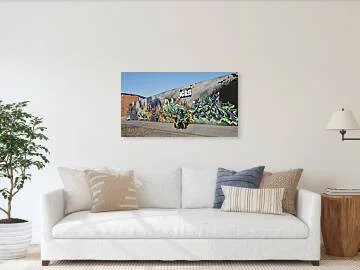 Two riders on a green motorcycle pass a long gray mural wall with gold, blue and violet-green lettering, shown in a room with sofa