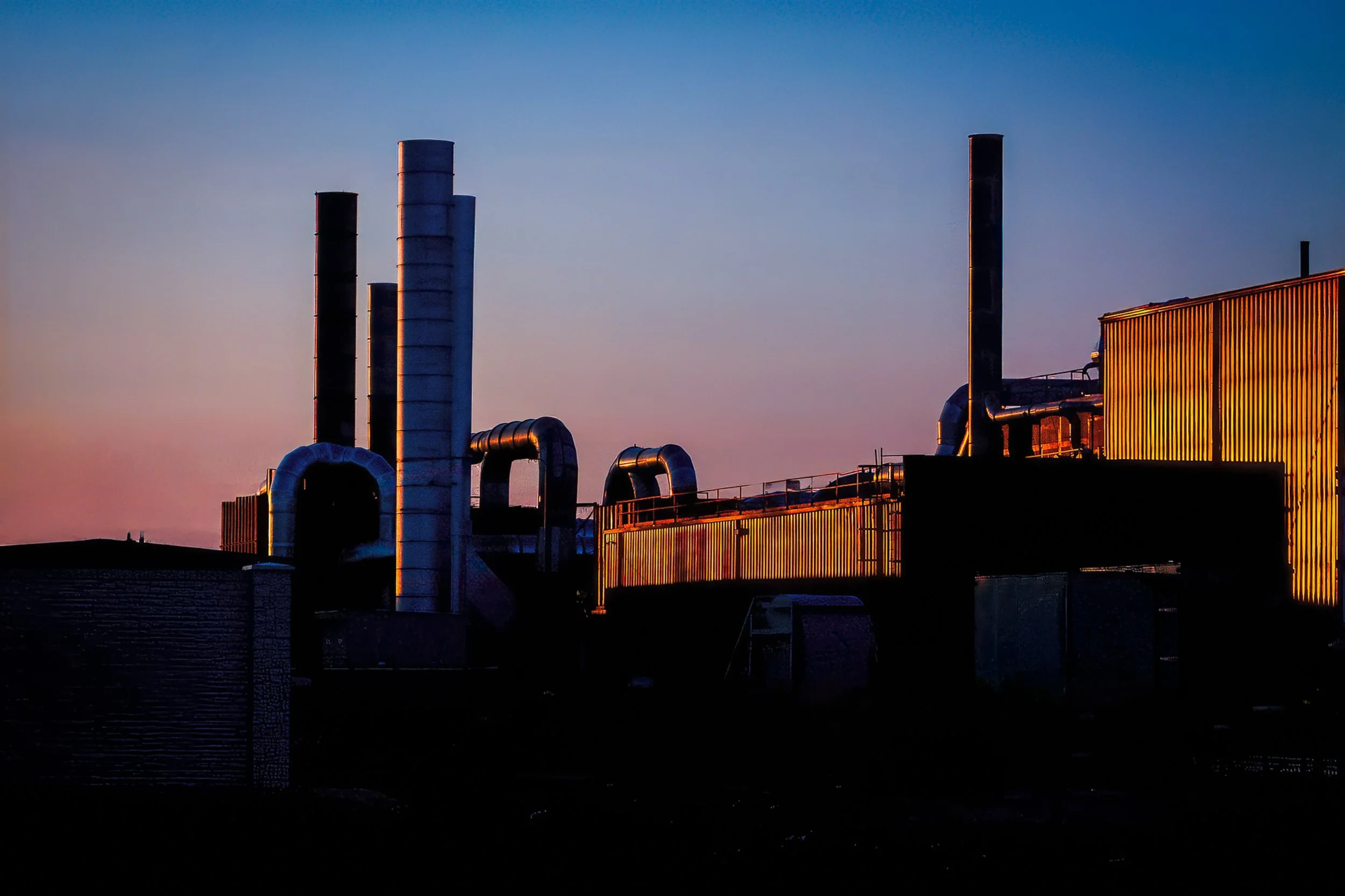 Last Light on the Line – industrial silhouette photograph of smokestacks and piping against a sunset gradient sky, one corrugated wall catching warm light