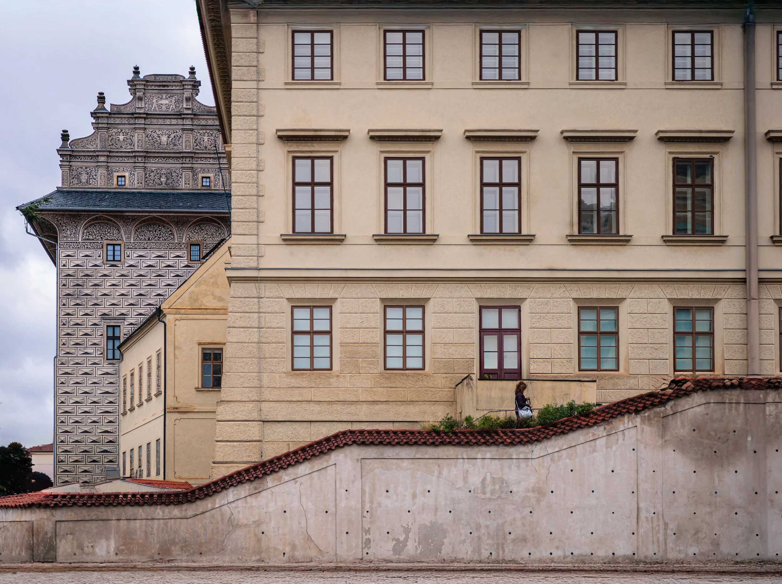 A lone woman walks along an elevated pathway between historic buildings in Prague beneath an ornate patterned tower.